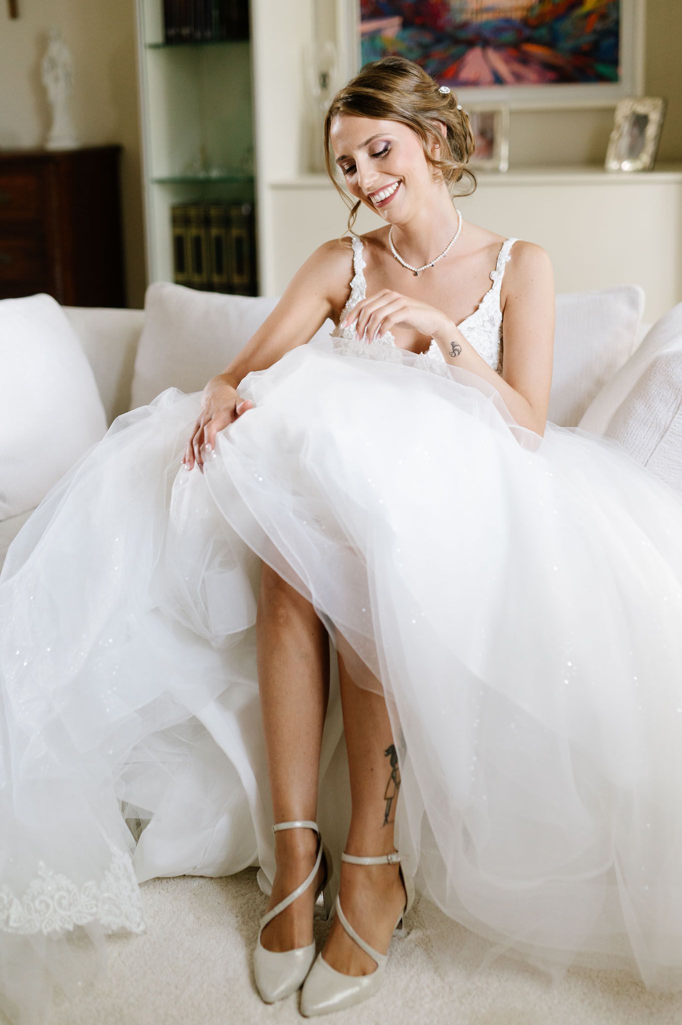 A bride in a flowing white gown sits elegantly on a white couch showing off her wedding shoes.