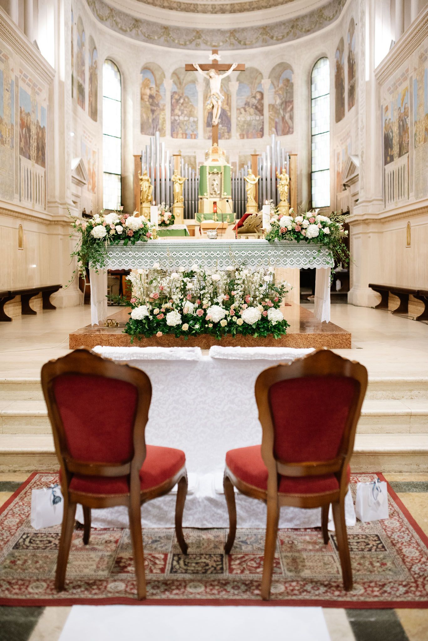 An ornate church altar decorated with white floral arrangements and two red velvet chairs.