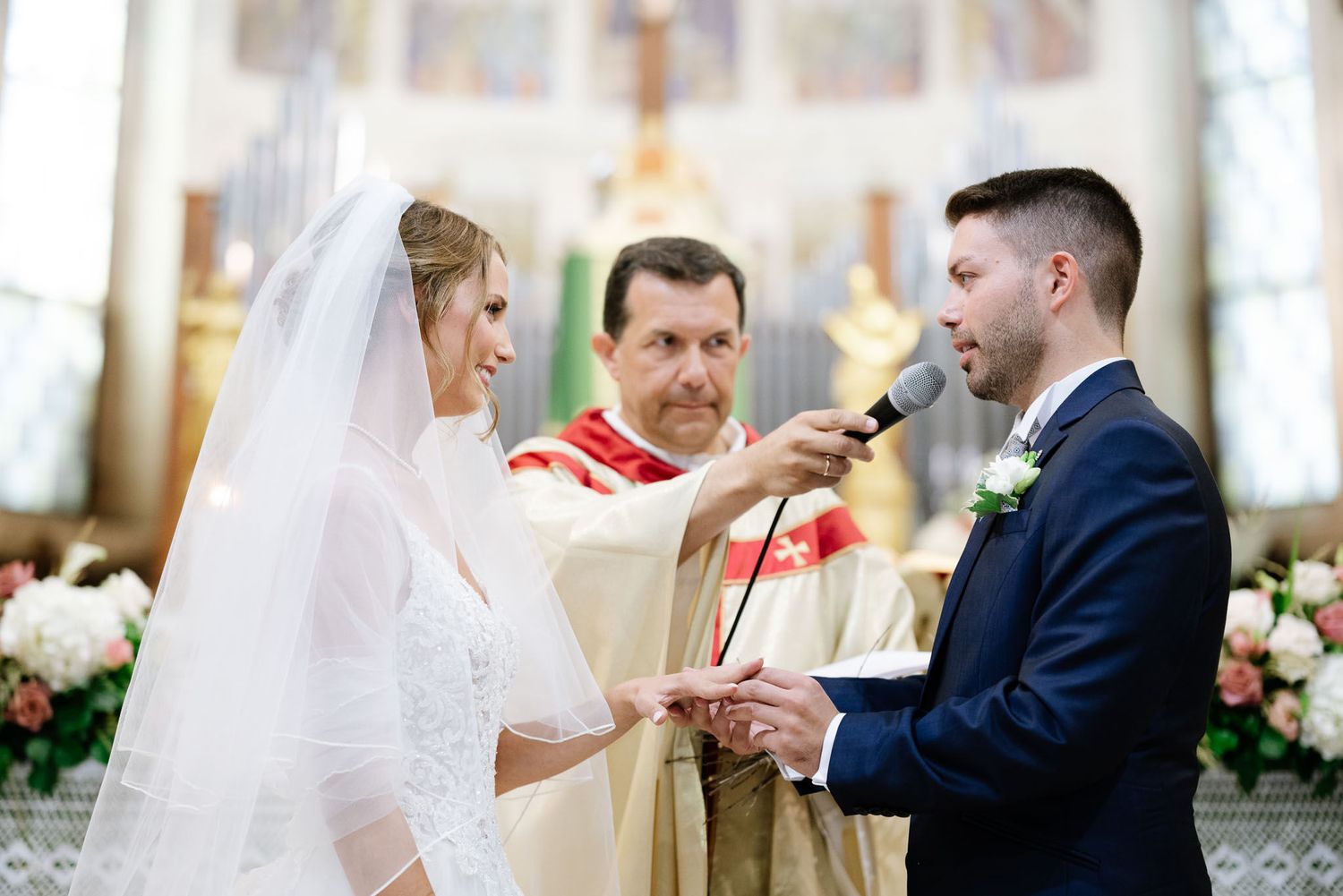 Traditional Catholic marriage ceremony with priest officiating as couple exchanges vows at decorated altar.