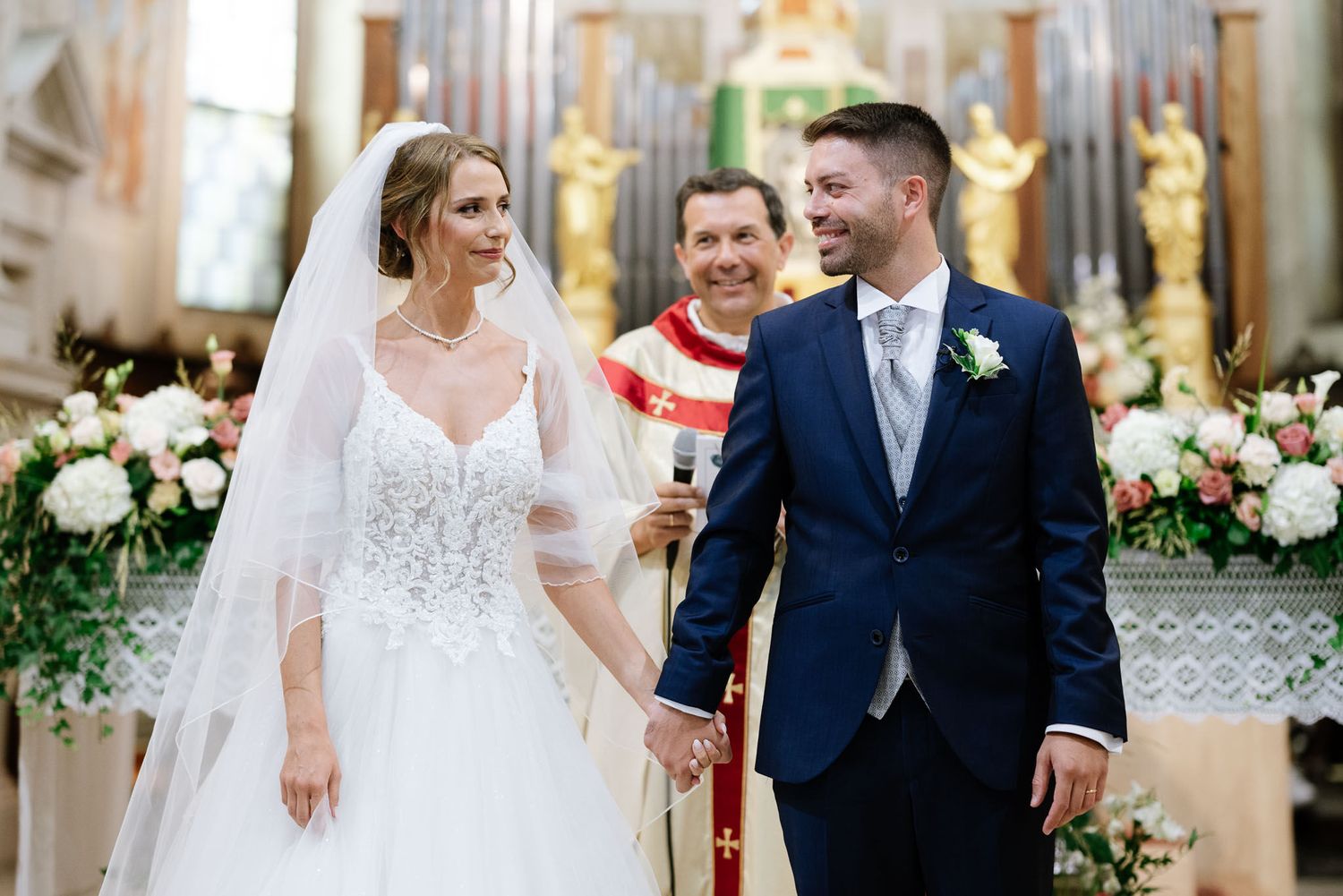 Joyful moment during Catholic wedding ceremony with couple and priest at flower-adorned altar.