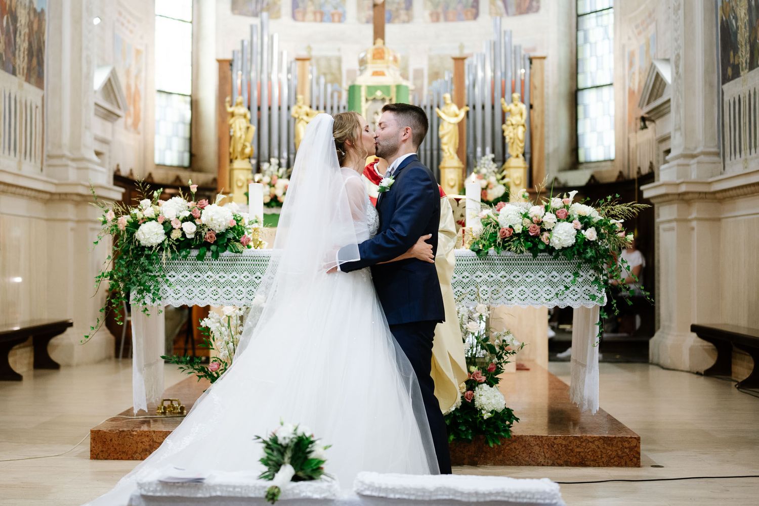 Romantic first kiss as newlyweds at altar surrounded by white floral arrangements in ornate church.