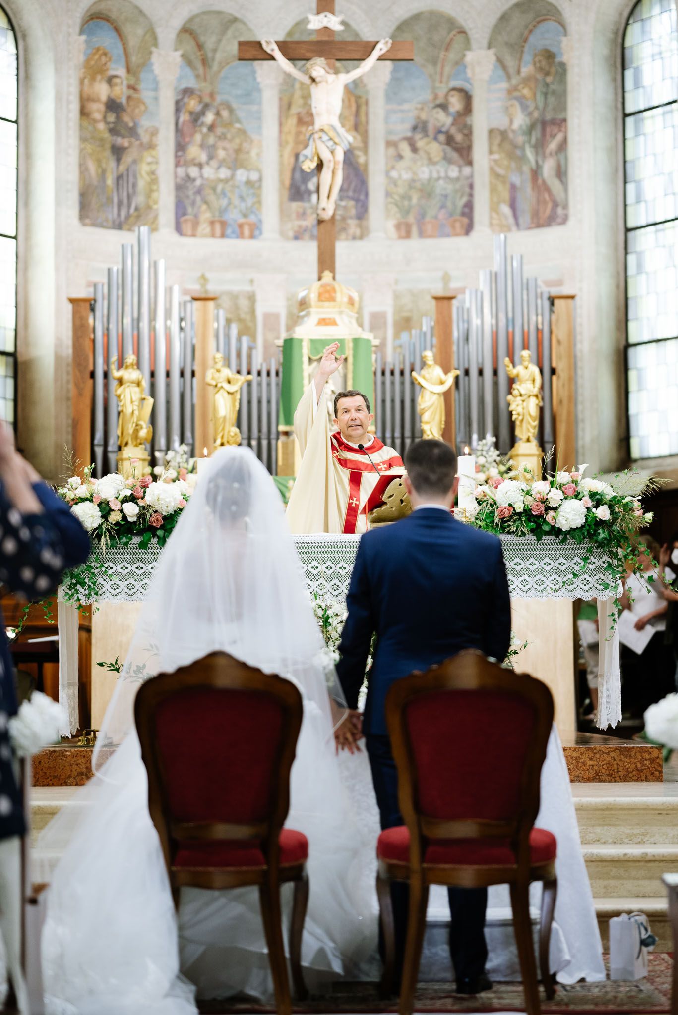 A couple sits at their Catholic wedding ceremony with an altar decorated with organ pipes and religious statuary.