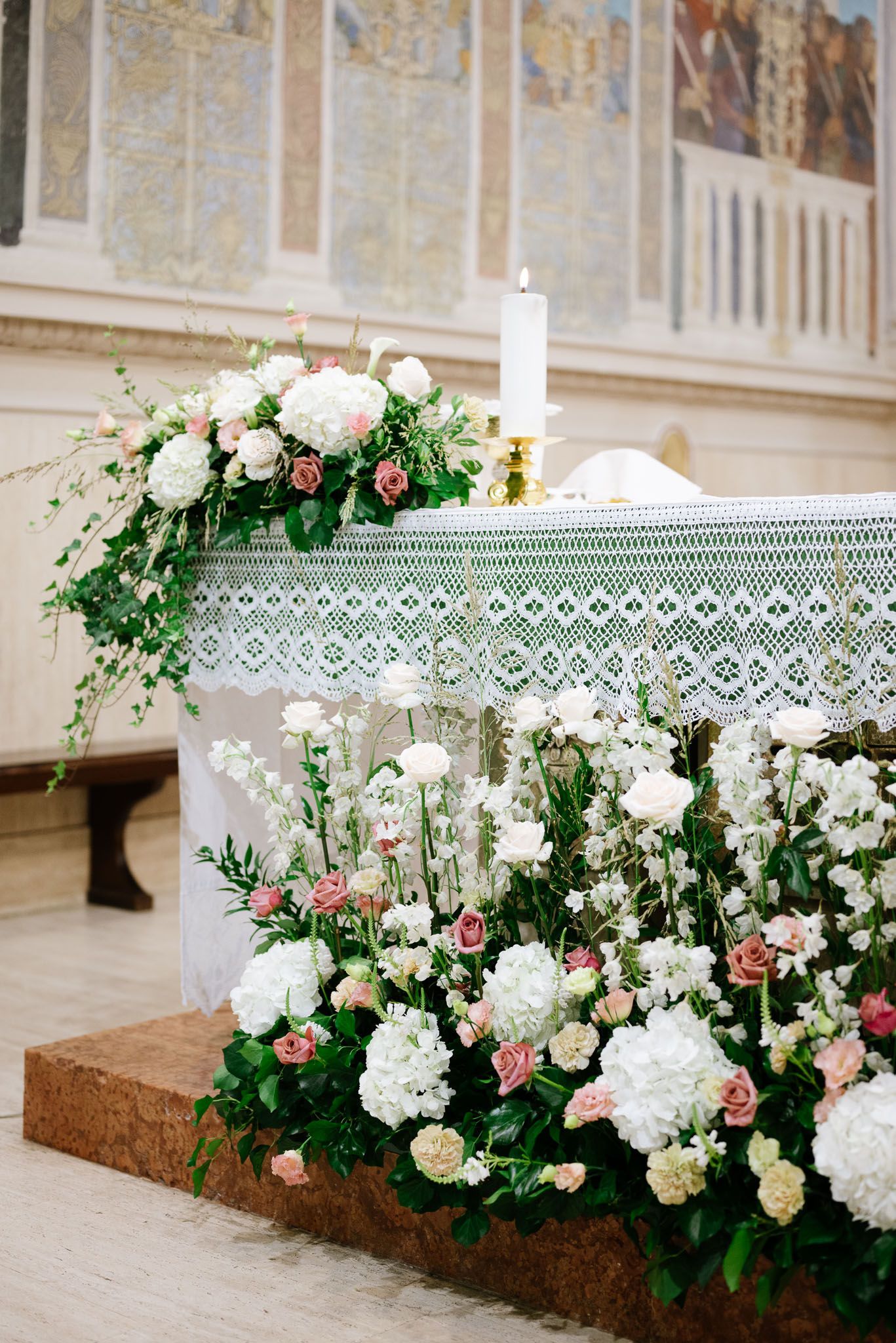 Elegant floral altar decoration with white lace tablecloth and lit candle in a church setting.