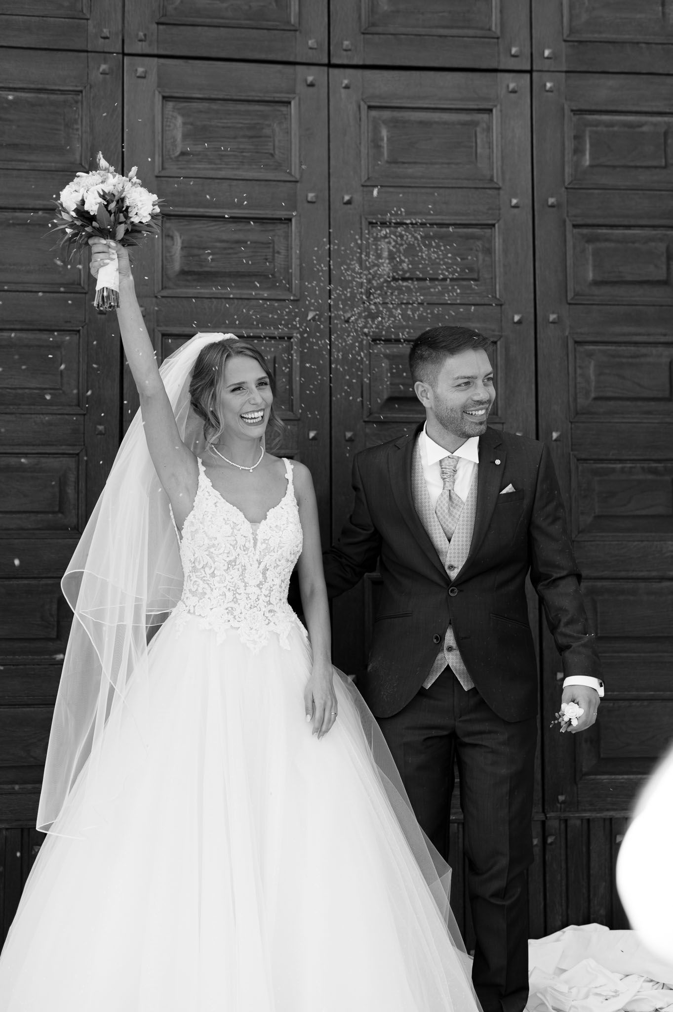 Newlyweds react joyfully to confetti shower against wooden church doors.
