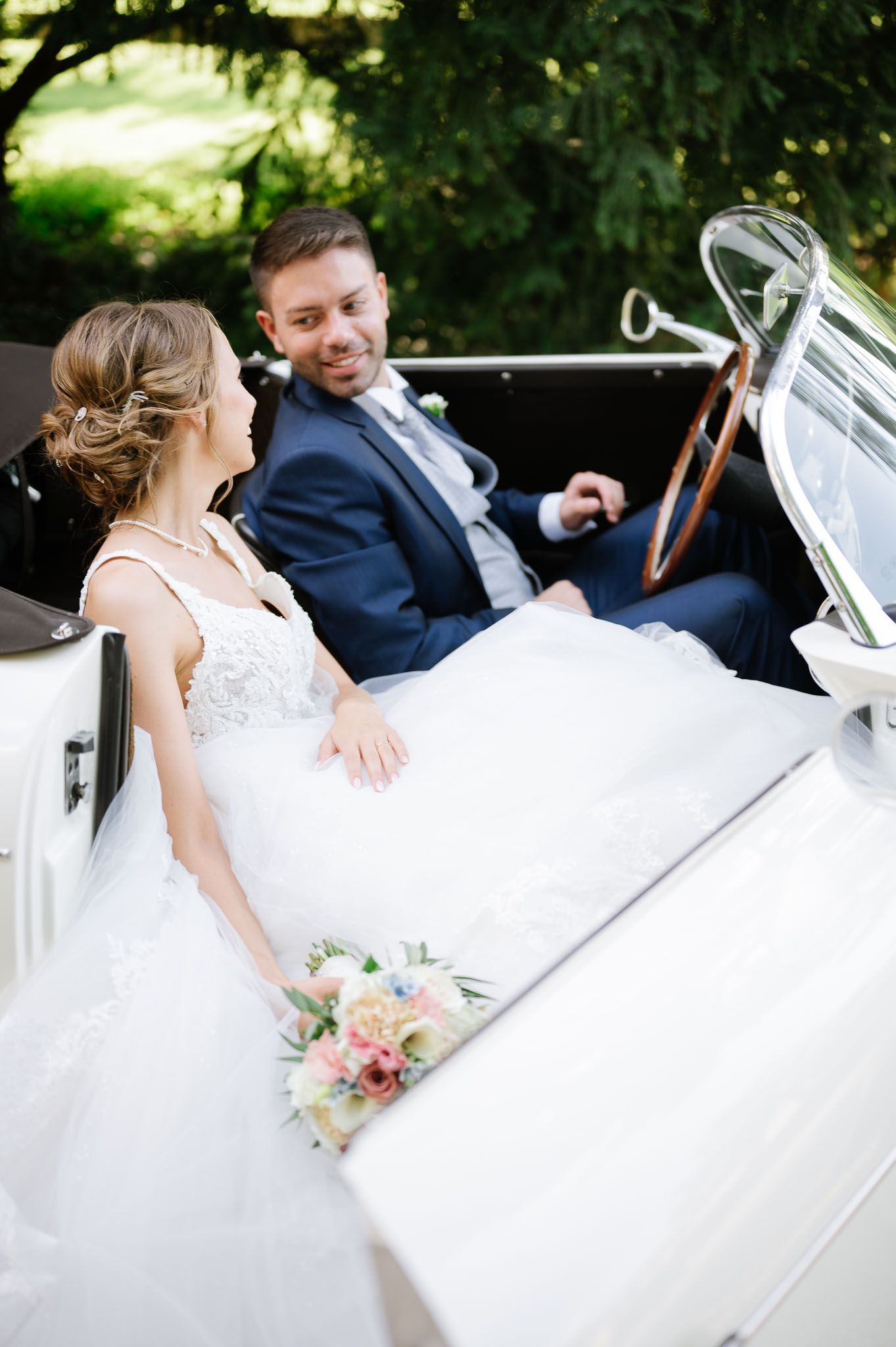 Newlyweds sitting together in a vintage white convertible with bridal bouquet.