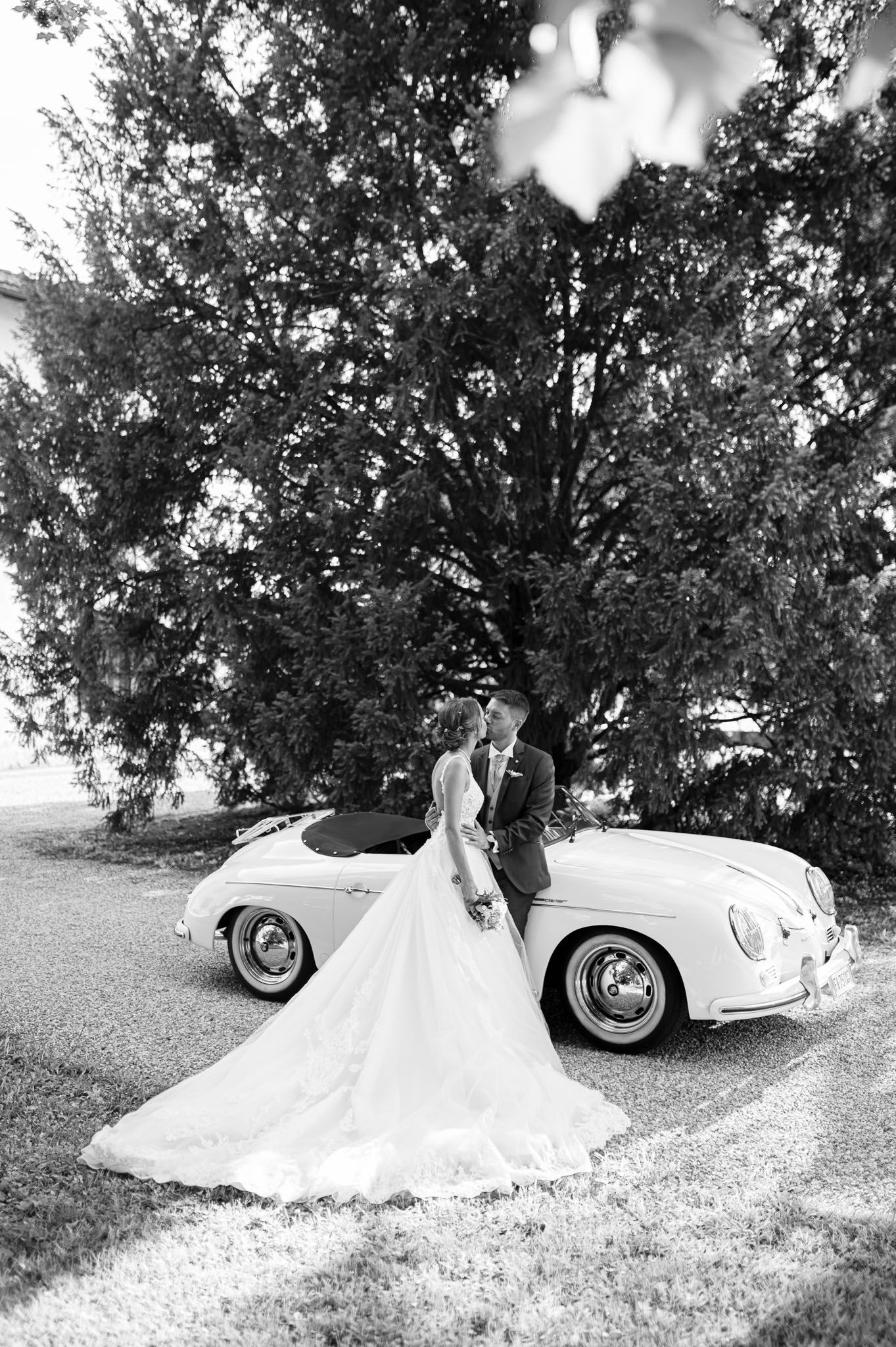Black and white wedding portrait with couple standing by classic Porsche on garden grounds.
