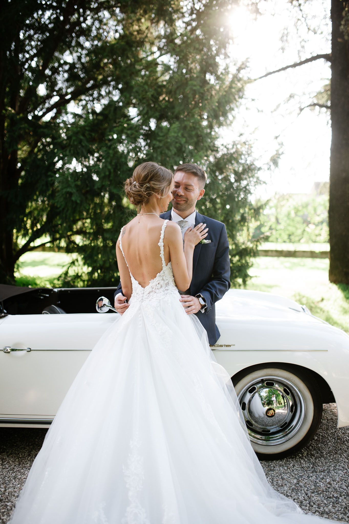Elegant wedding couple poses beside classic white convertible on gravel path.