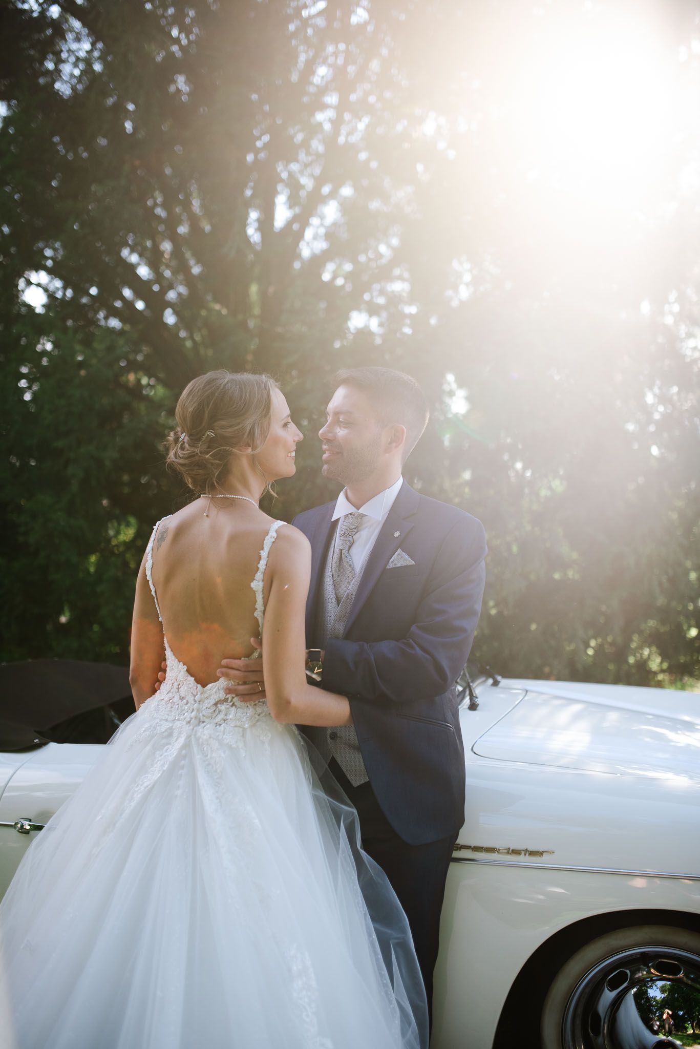 Sunlit wedding portrait of couple embracing by vintage car.