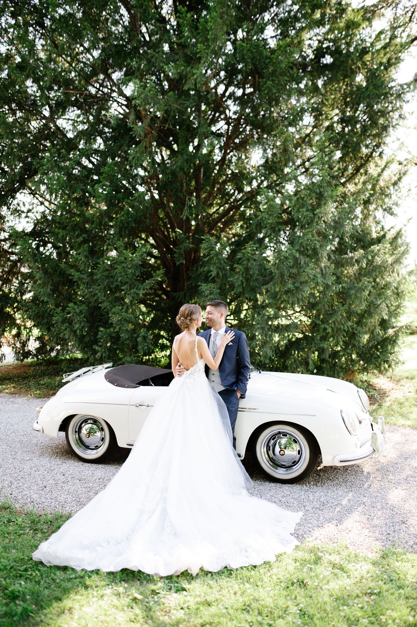 Romantic wedding portrait with couple standing by vintage Porsche against towering trees.