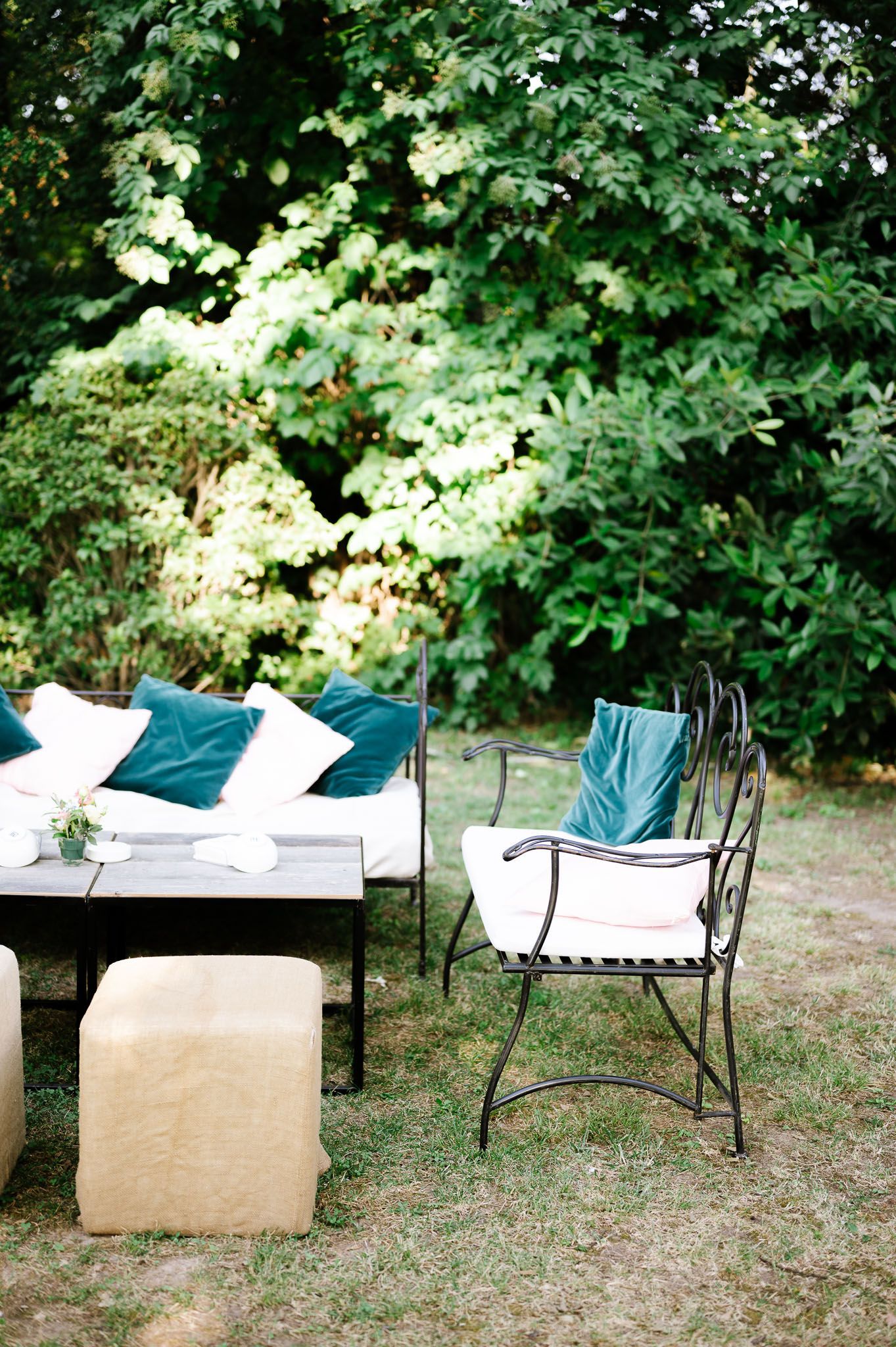 Outdoor seating area with white bench, teal pillows and wrought iron chairs in a garden setting.