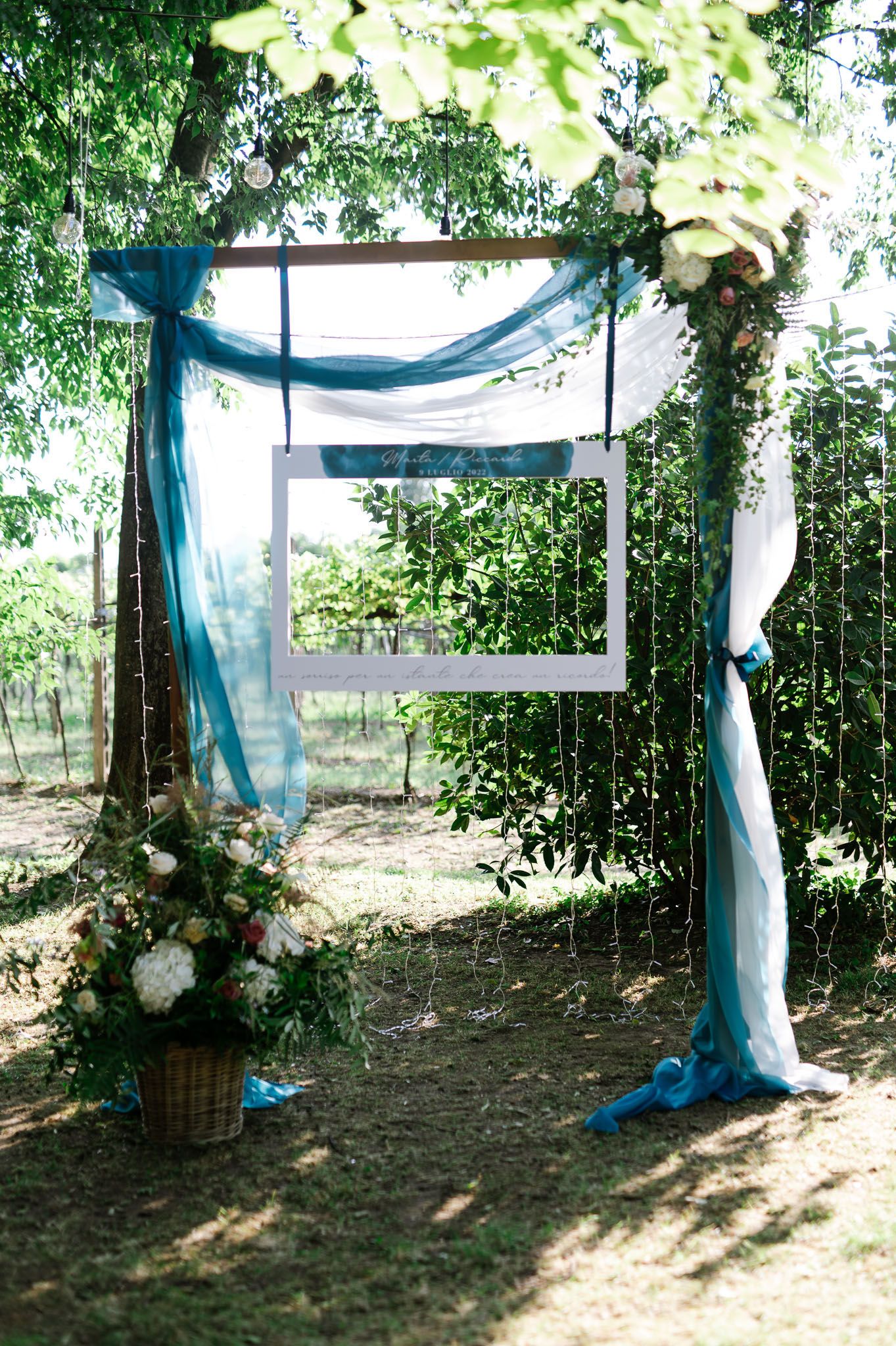 Wedding arch decorated with flowing blue fabric, white frame, and floral arrangements in a wooded setting.