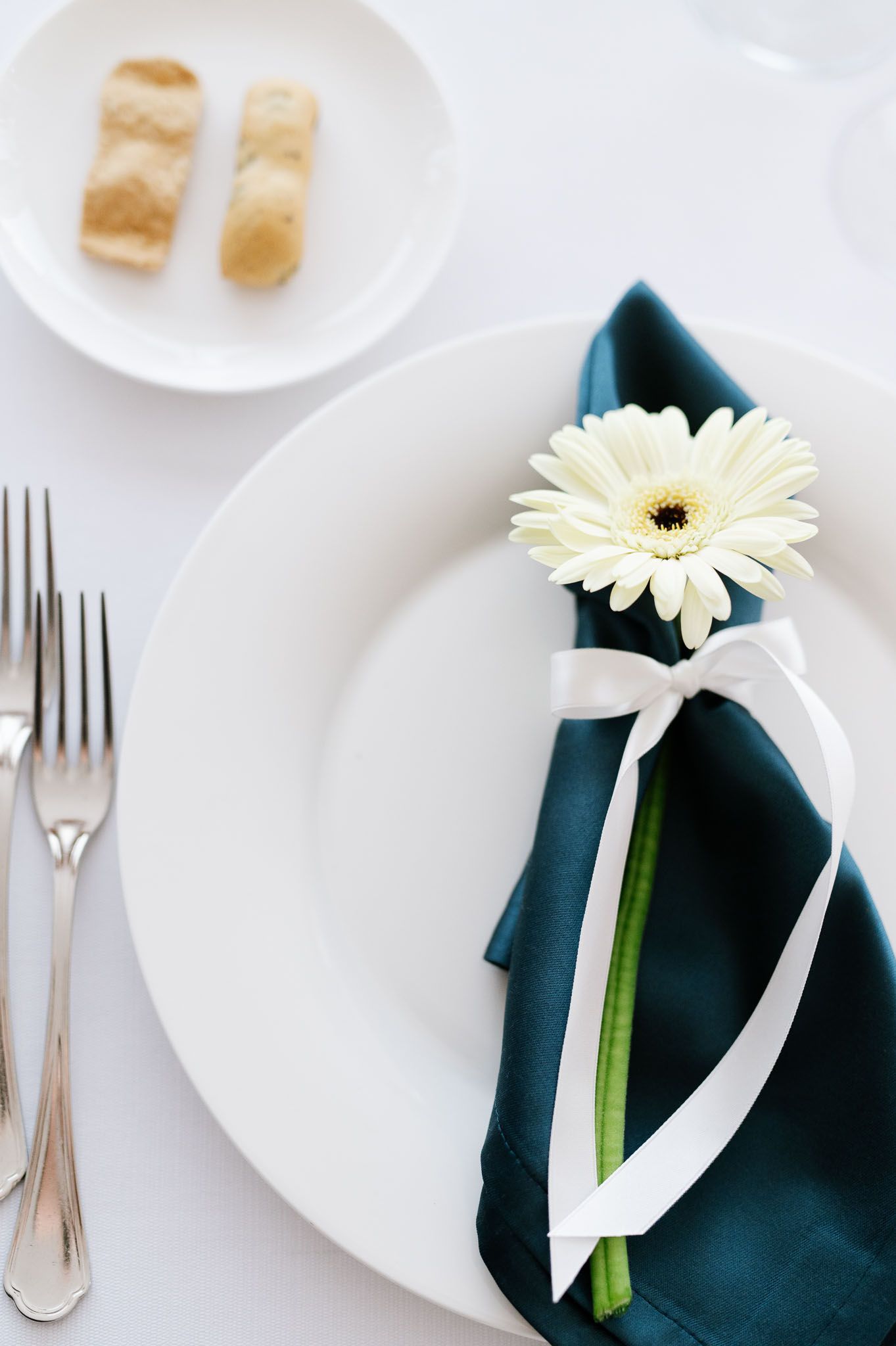 Place setting with white plate, teal napkin and white gerbera daisy tied with ribbon.