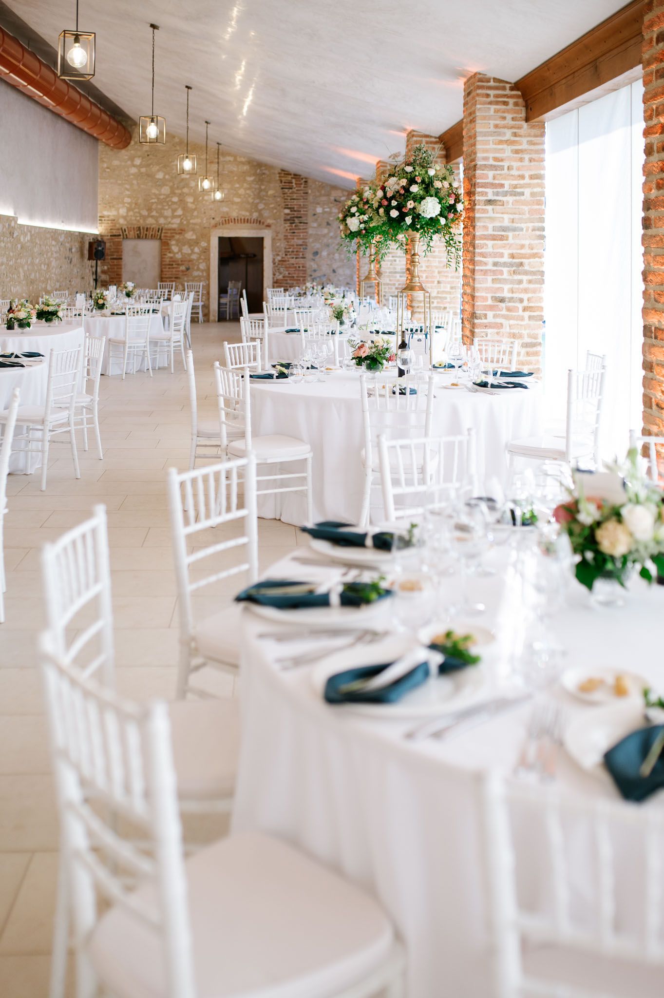 Elegant wedding reception venue with white tables, chairs and floral centerpieces in a brick-walled room.
