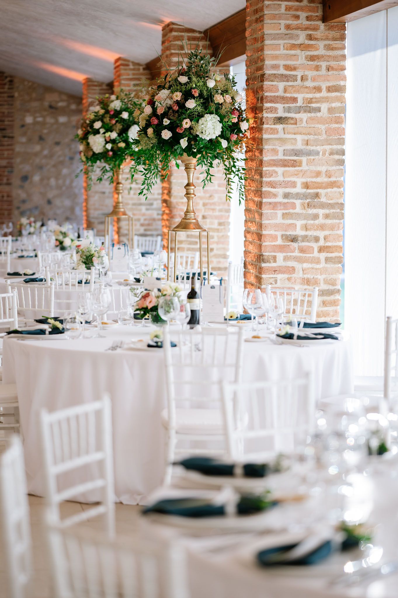 Wedding reception tables with tall floral centerpieces and white chiavari chairs in an elegant brick venue.