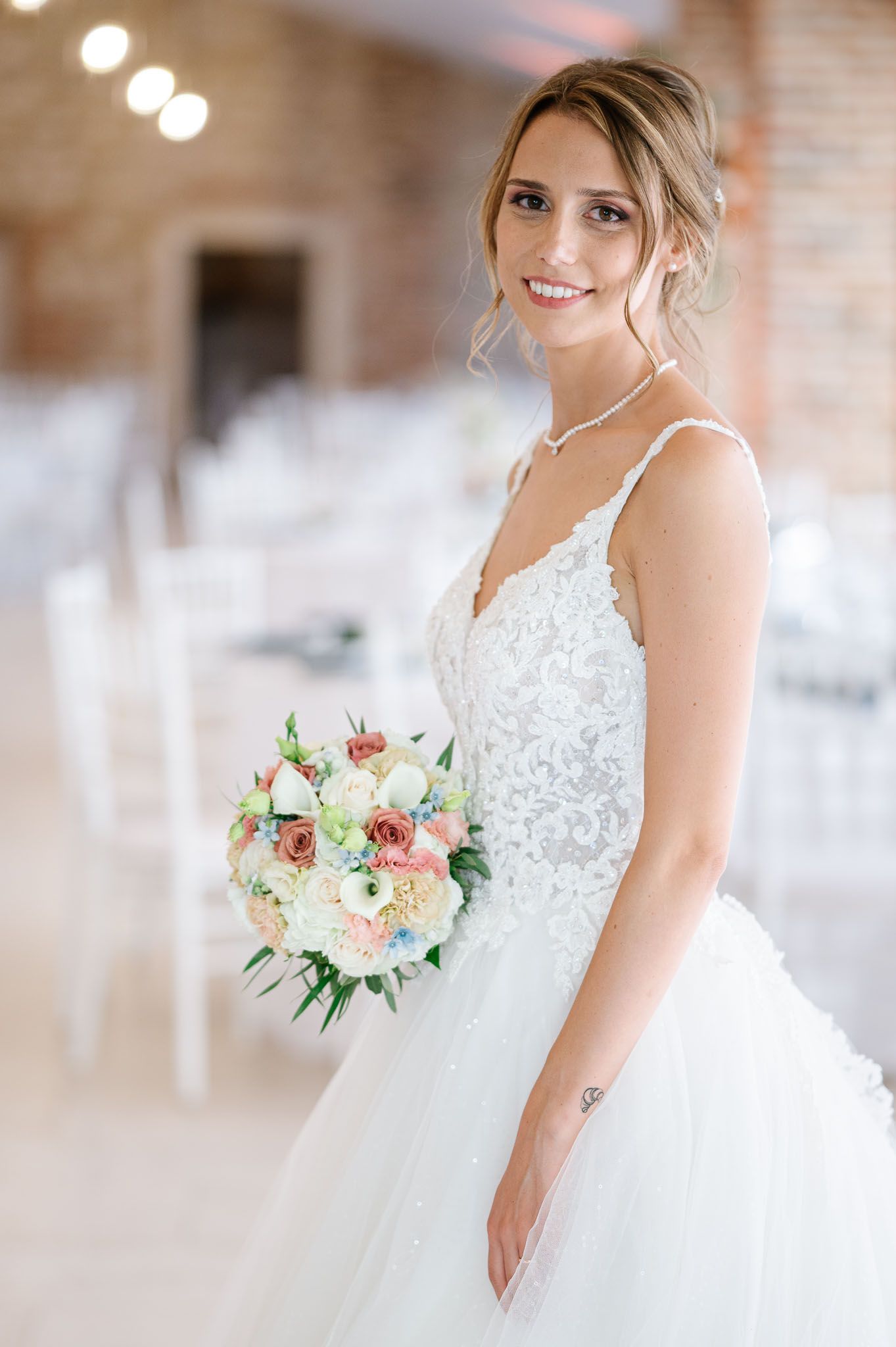 White lace wedding dress with tulle skirt and delicate floral bouquet in a rustic brick venue.
