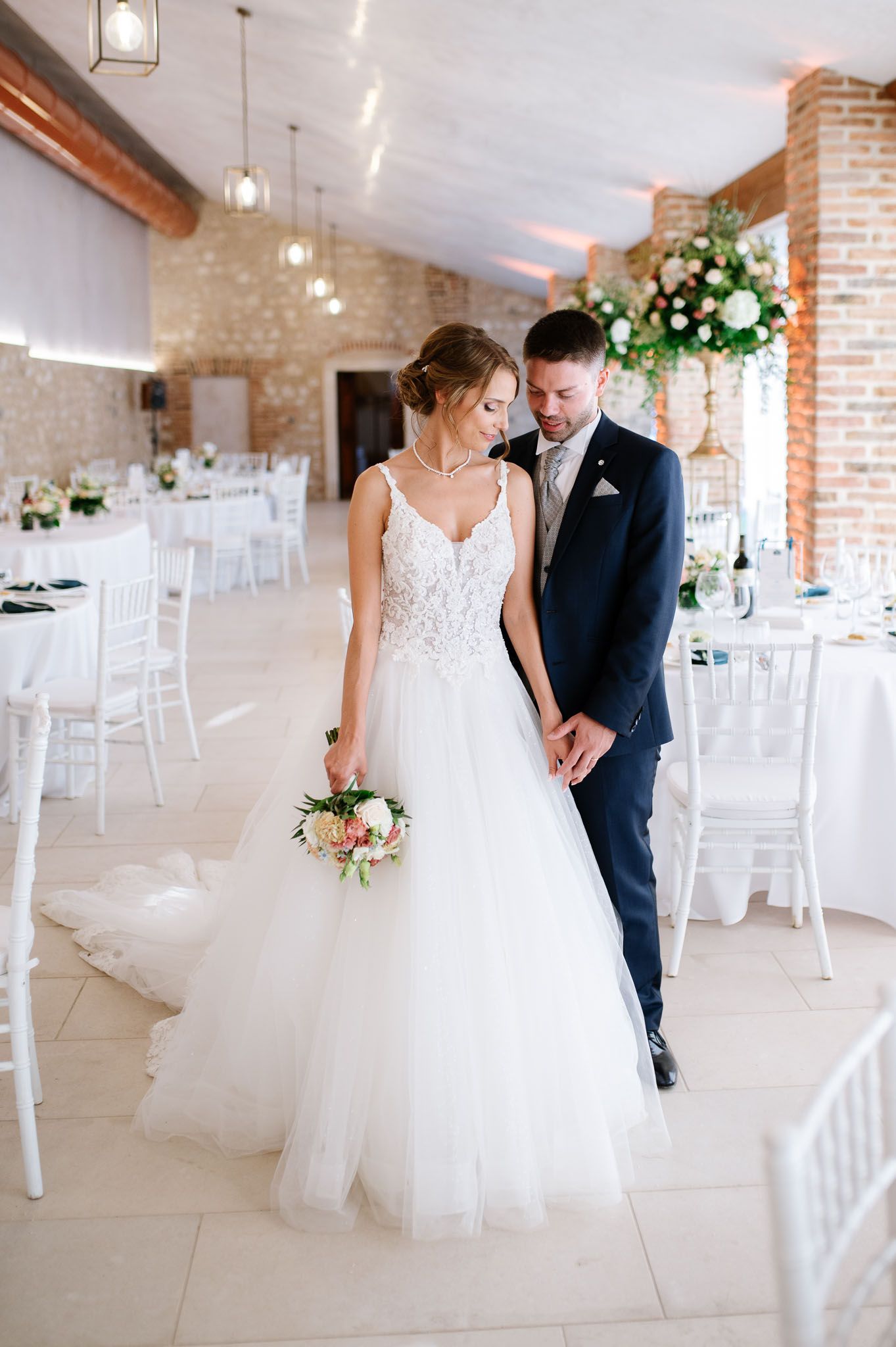 Newlyweds share an intimate moment in a rustic brick reception hall with floral decorations.