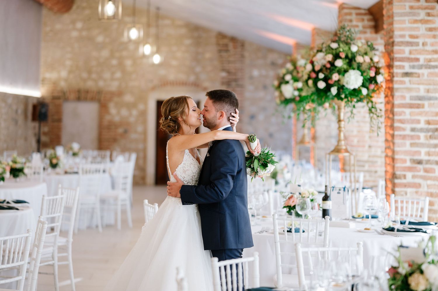 Romantic embrace in a reception hall decorated with hanging lights and floral arrangements.