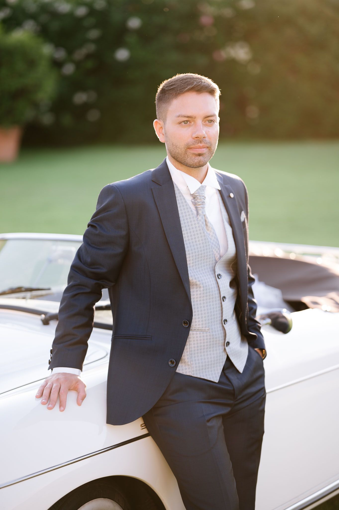 Groom in navy suit with grey waistcoat leans against white car outdoors.