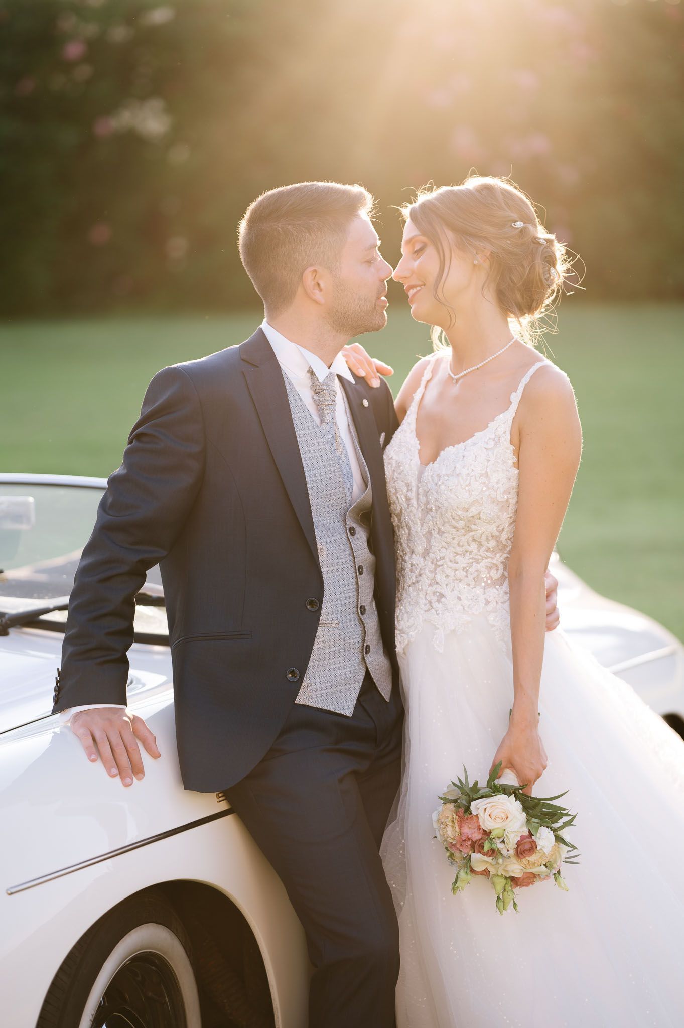 Newlyweds share an intimate moment against a classic car in golden sunset light.