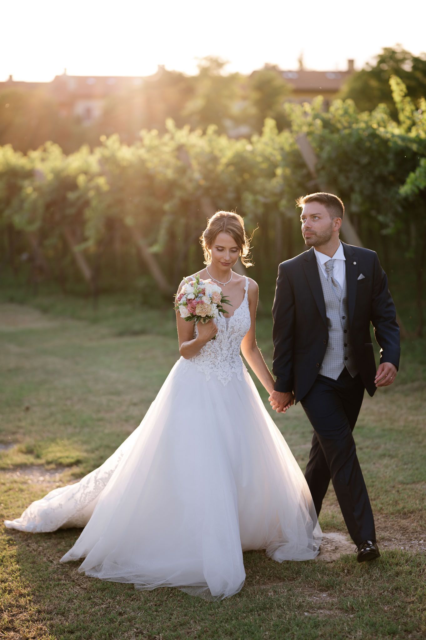 A newlywed couple walks hand in hand through a vineyard at sunset.