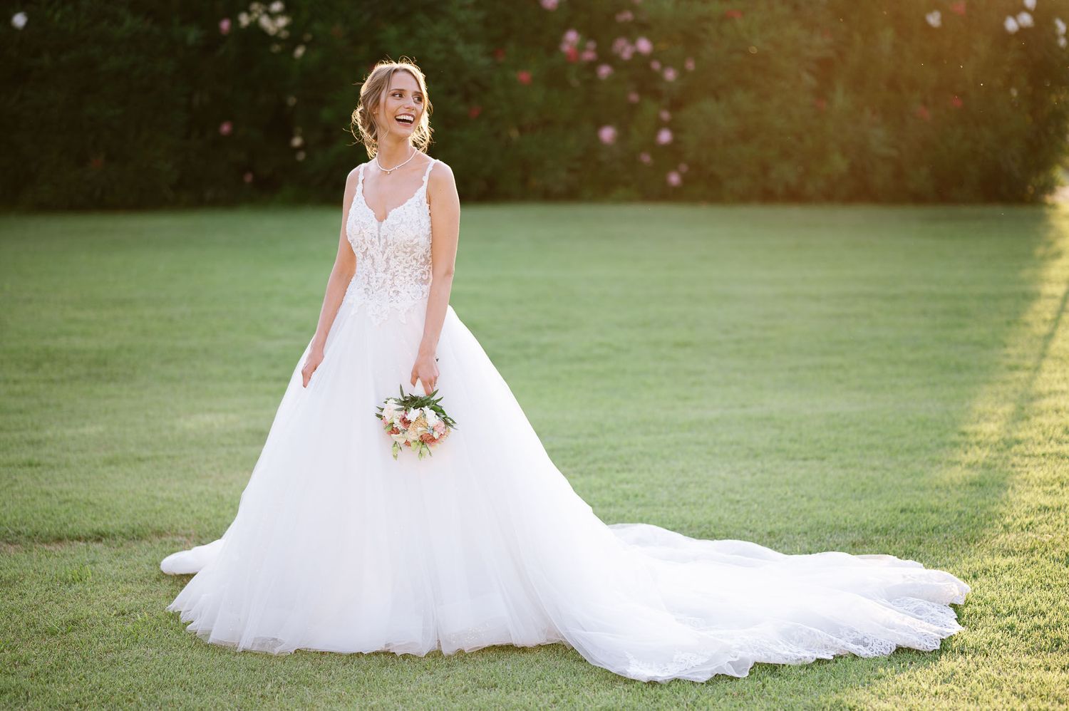A bride in a white lace ballgown poses on a sunlit lawn while holding a delicate bouquet.