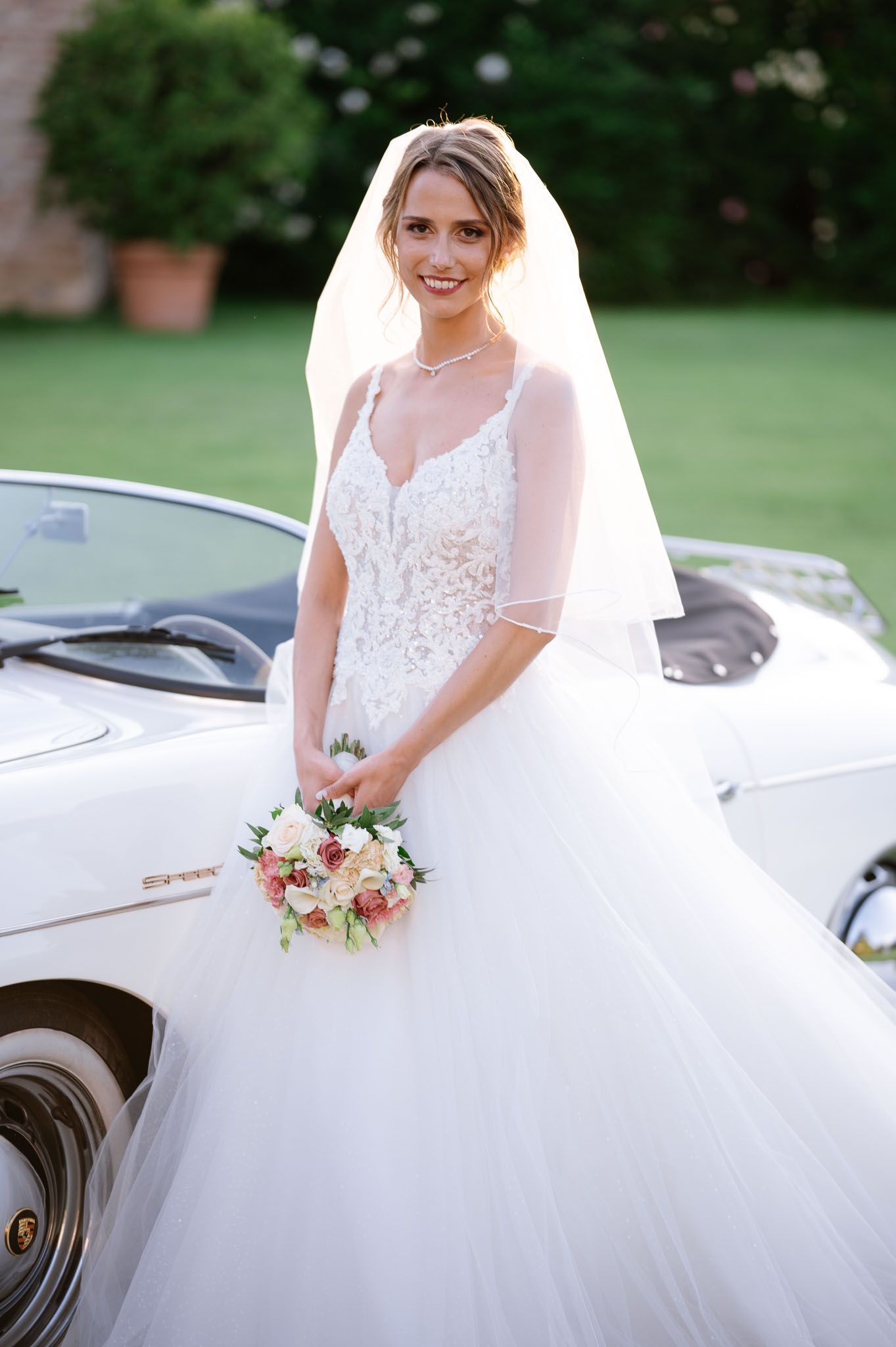 A bride poses beside a vintage car wearing a flowing tulle gown and holding a pink bouquet.