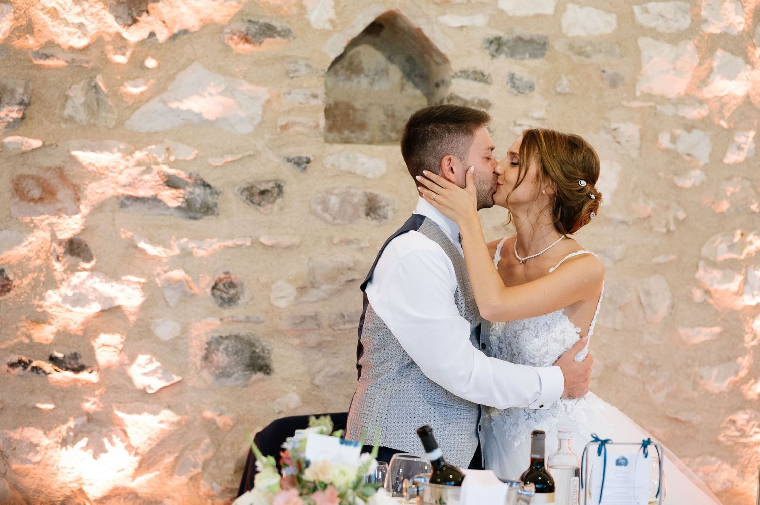 A romantic wedding kiss captured against a rustic stone wall with wedding cake in foreground.