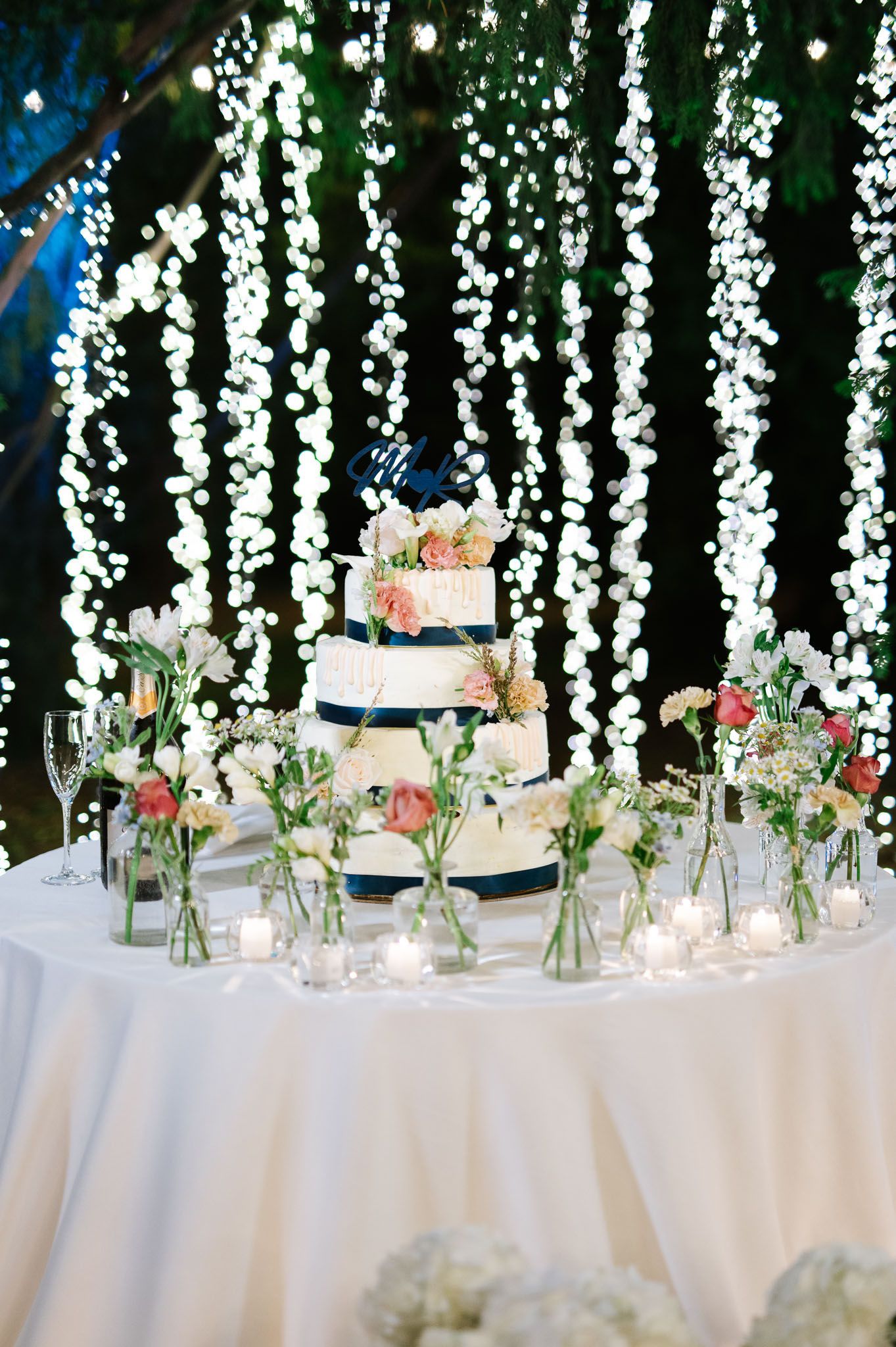 A three-tiered wedding cake displayed under cascading fairy lights with floral arrangements.
