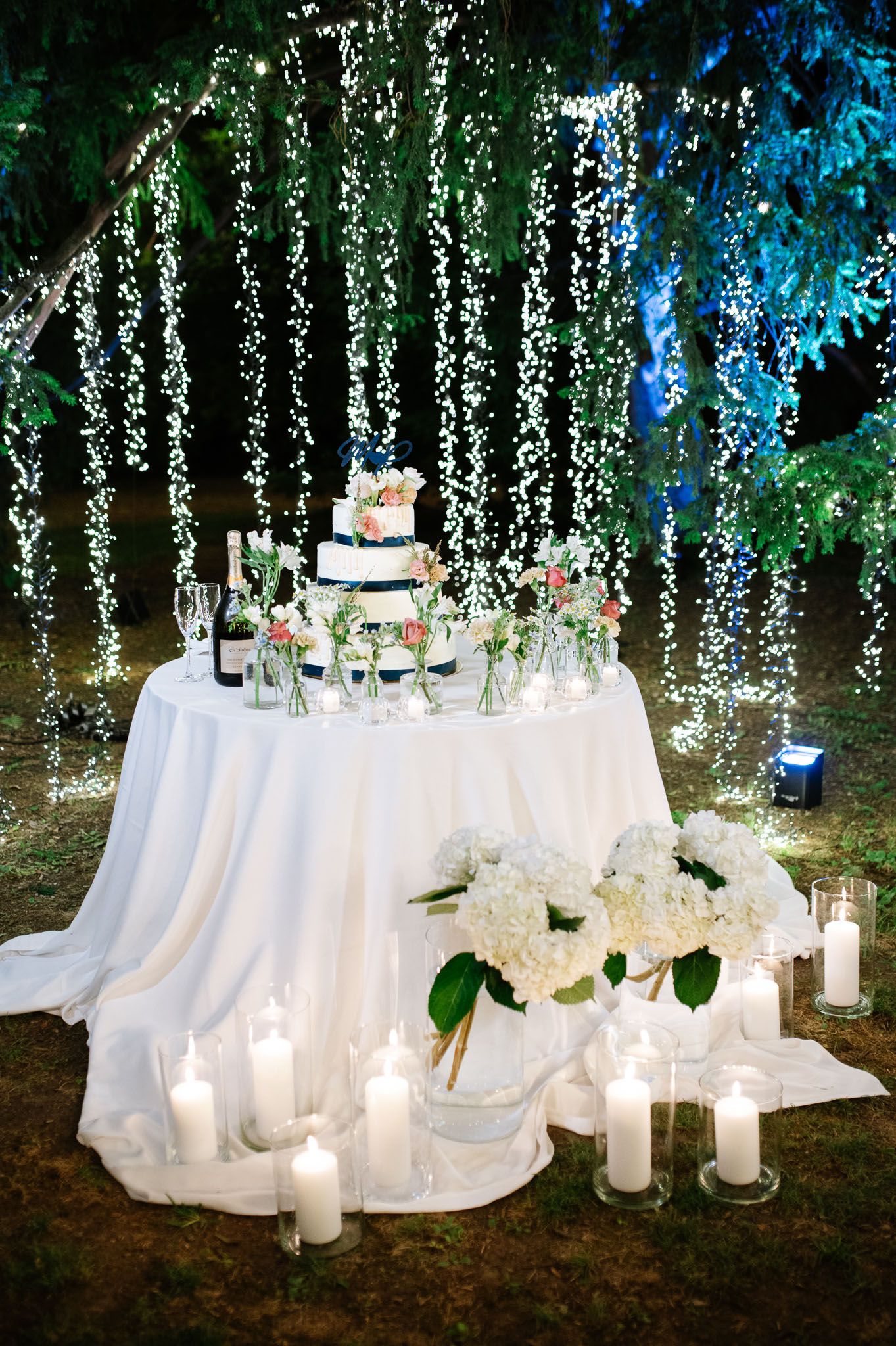 A wedding cake and candle display beneath hanging string lights creates a magical evening ambiance.