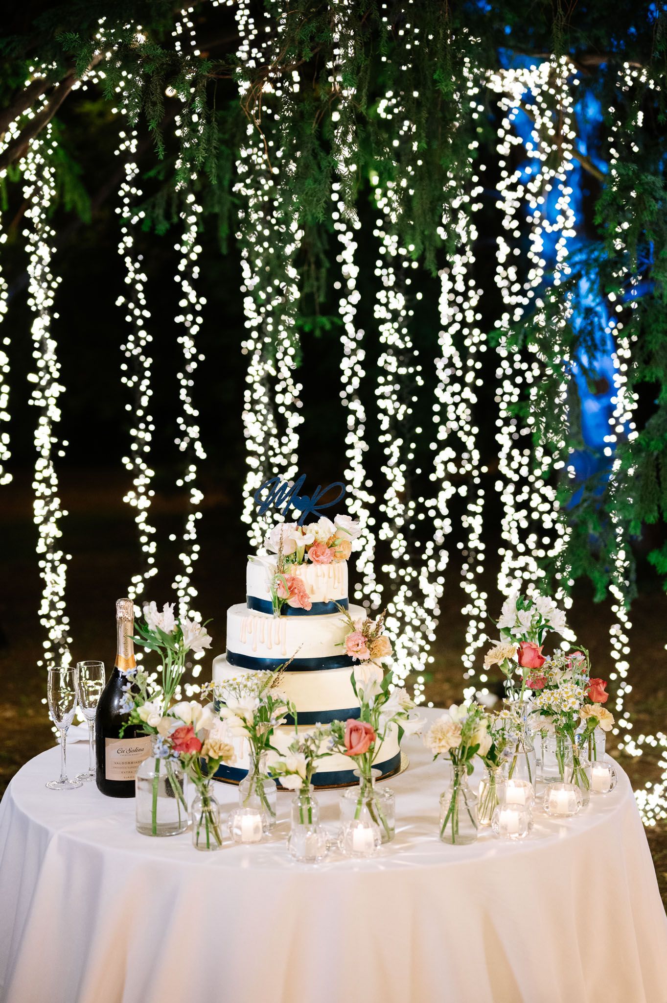 Elegant wedding cake table adorned with cascading string lights and delicate floral arrangements against a dark backdrop.