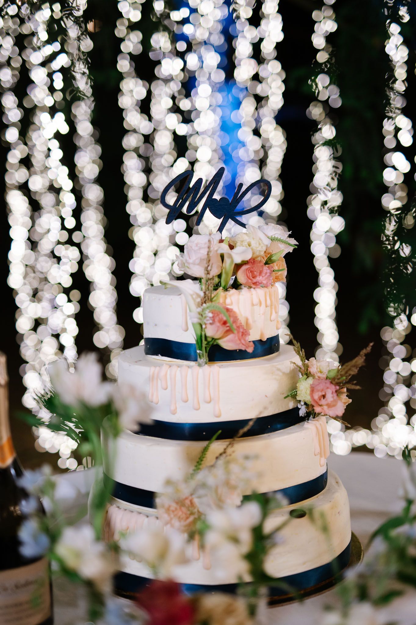 Four-tier white wedding cake with navy ribbons and pink floral accents under twinkling string lights.
