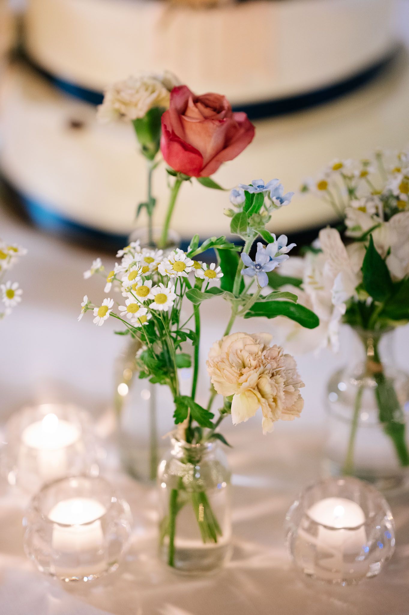 Small glass vases with pink roses and white flowers alongside tea lights on a wedding table.