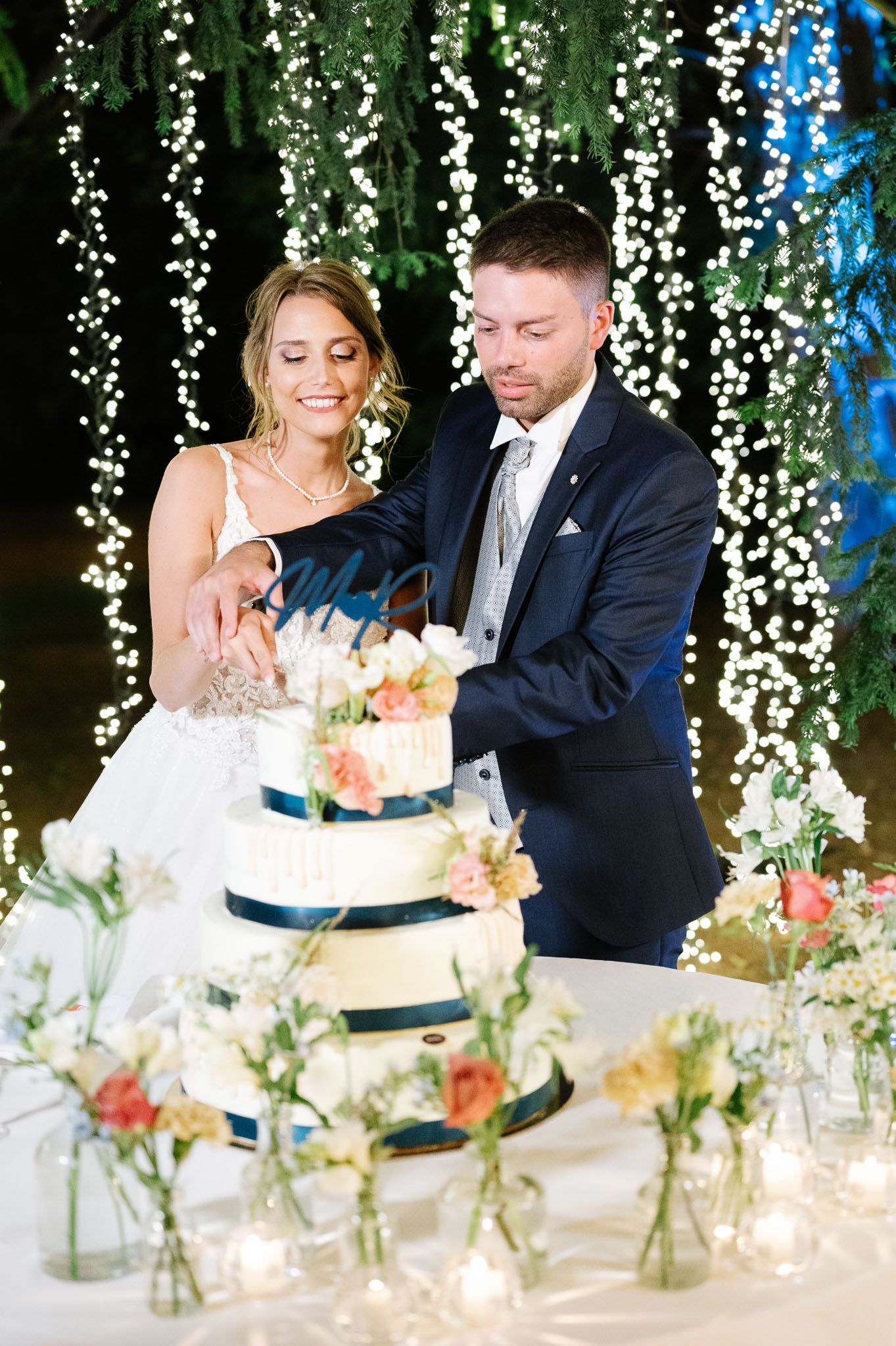Couple cutting their elegant four-tier wedding cake decorated with navy ribbons and fresh flowers.
