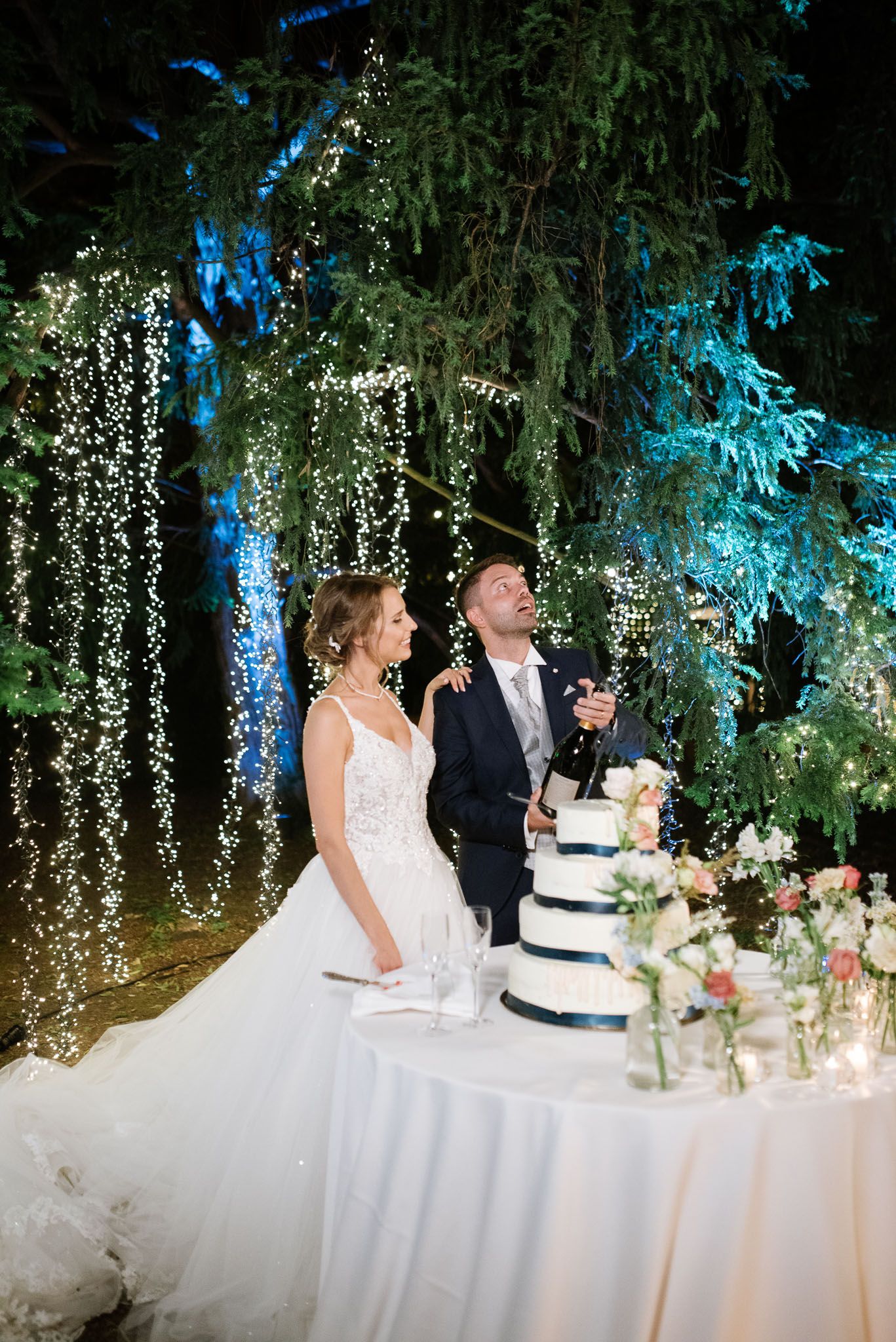 Wedding cake cutting scene under a magical canopy of hanging fairy lights and blue-lit trees.