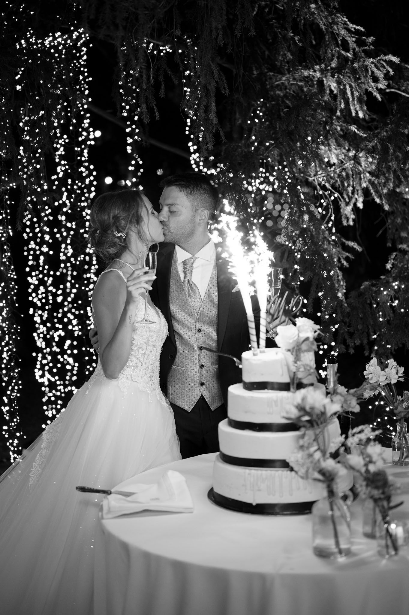 Black and white wedding moment captured under a cascade of twinkling lights with tiered cake.