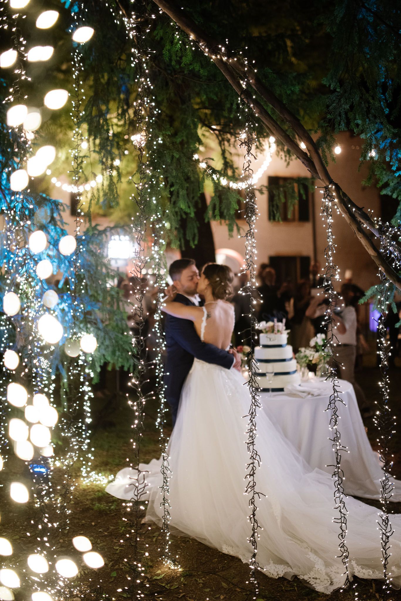 Enchanting wedding scene with hanging lights creating bokeh effect around couple and cake table.