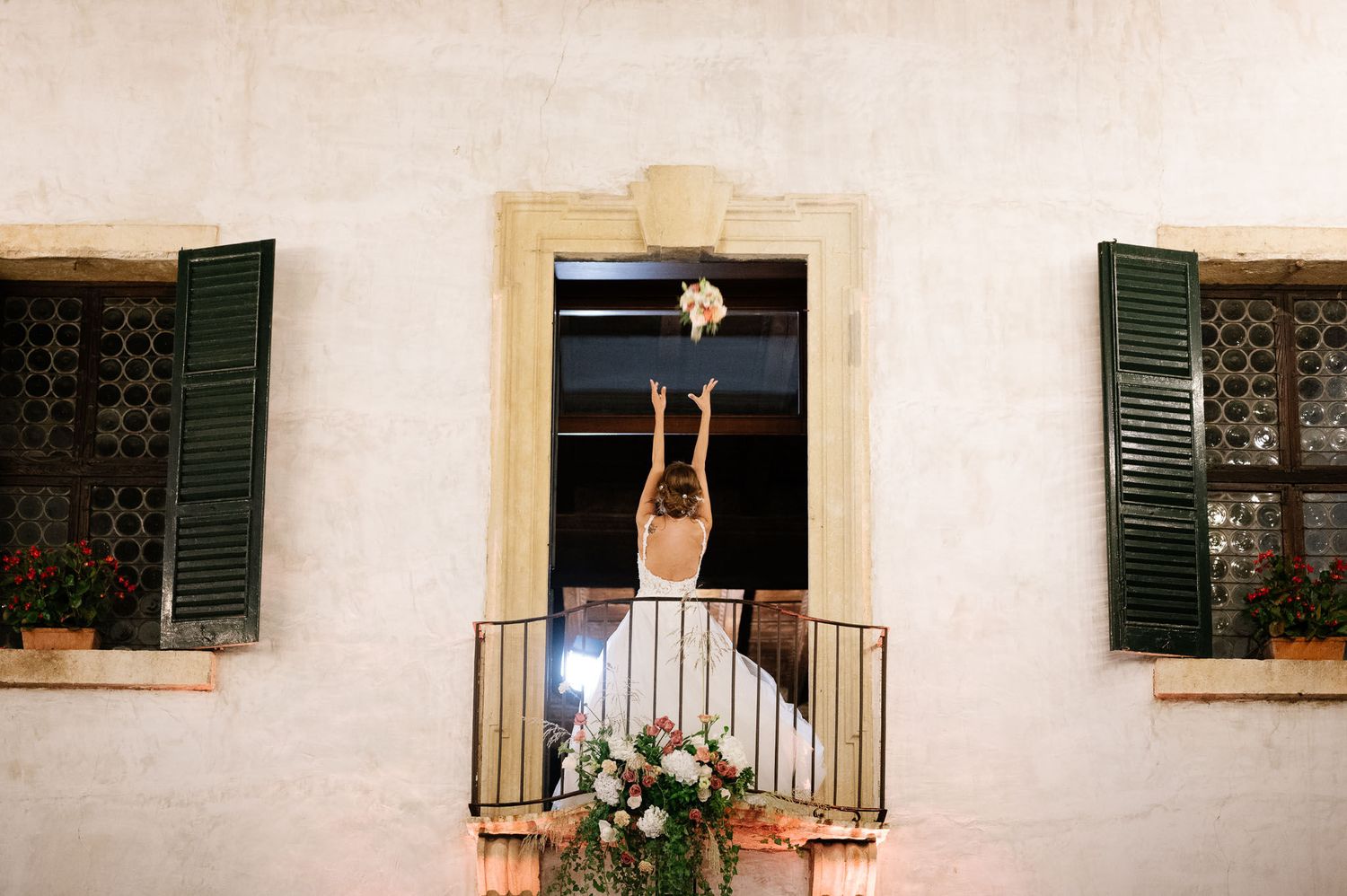 Wedding celebration scene with figure reaching from a flower-adorned balcony of a Mediterranean villa.