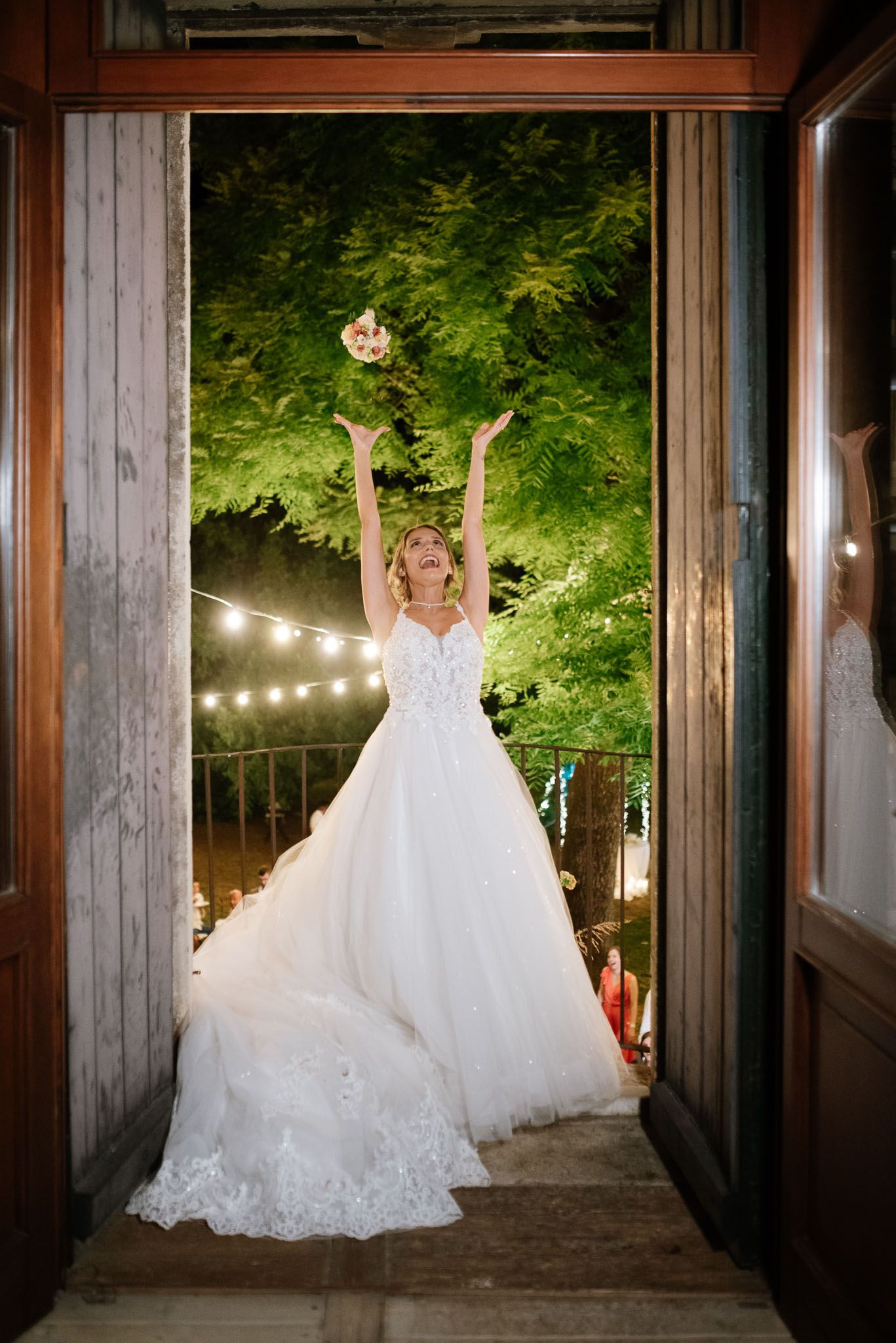 Bride in flowing white gown twirls joyfully beneath string lights in an elegant doorway.
