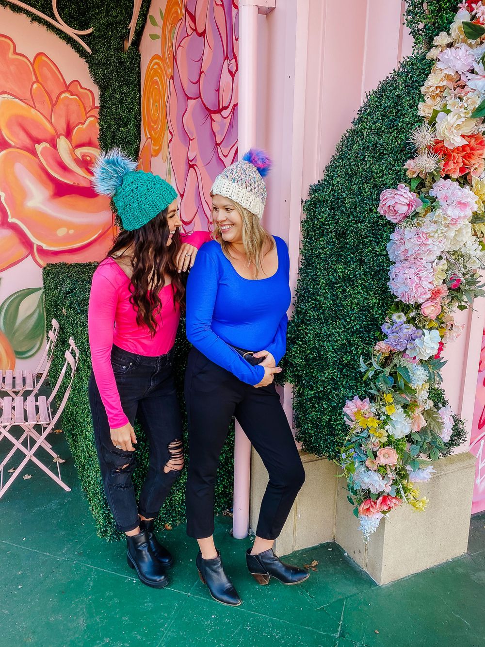 Two people in winter hats posing against a floral wall with decorative greenery.