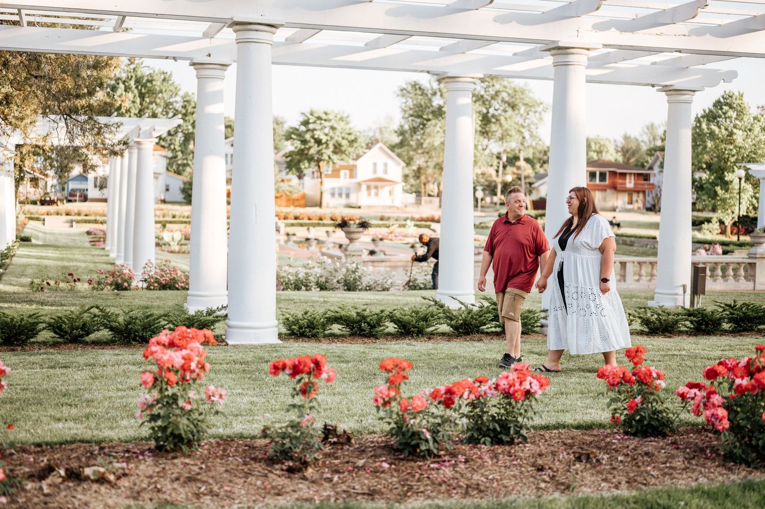 Couple walking through garden with white pillars and red flowers in foreground.