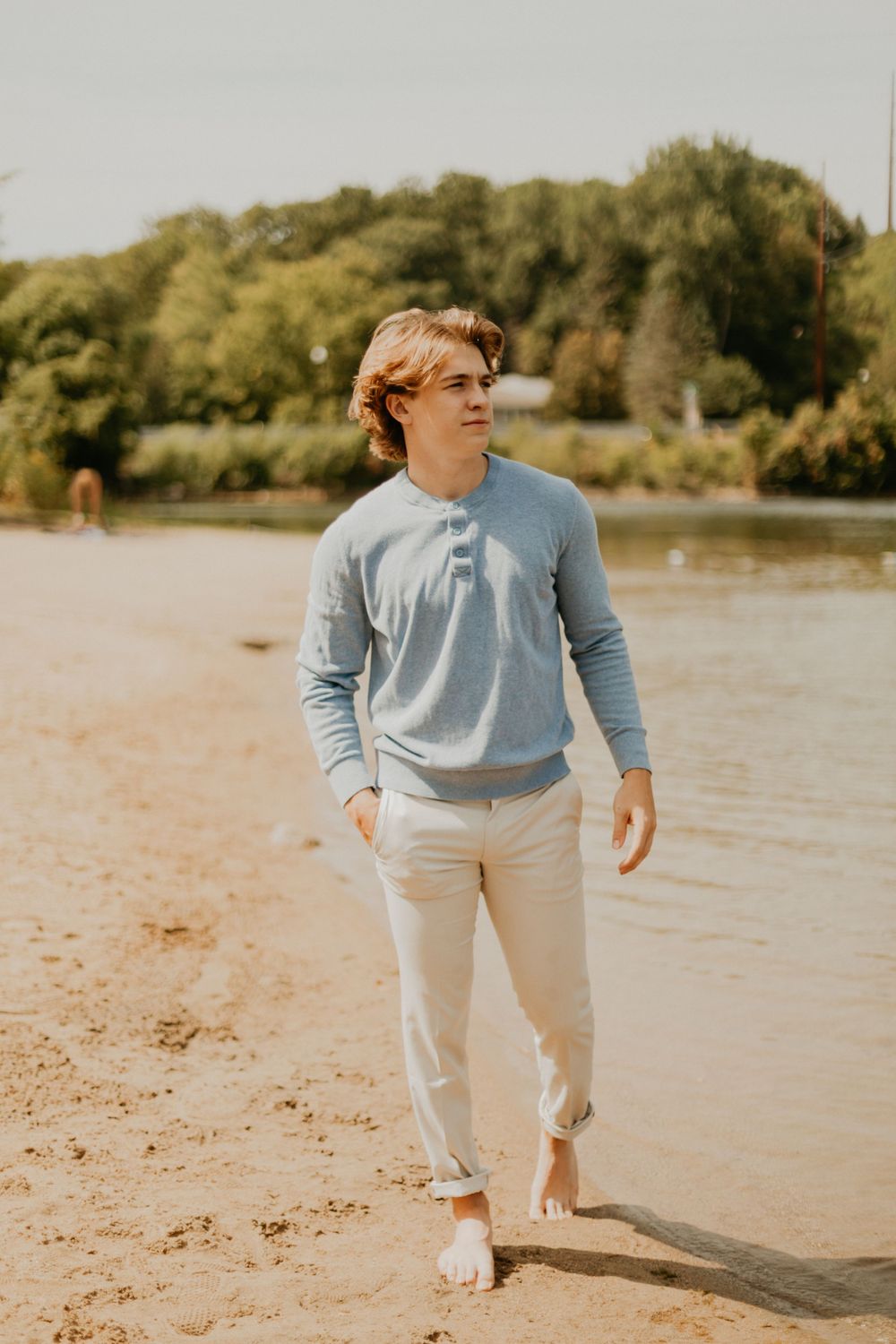 A person in light blue and white casual wear walks along a sunny beach shoreline.