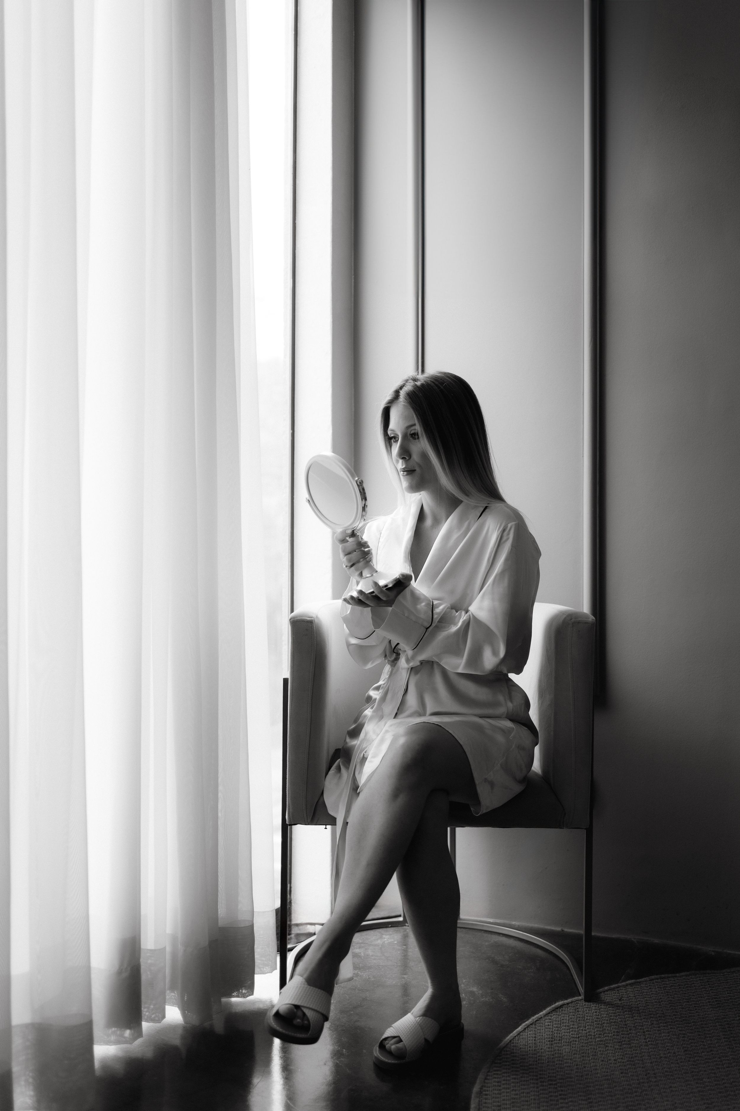 A black and white photo of someone in a robe sitting by a window holding a mirror.