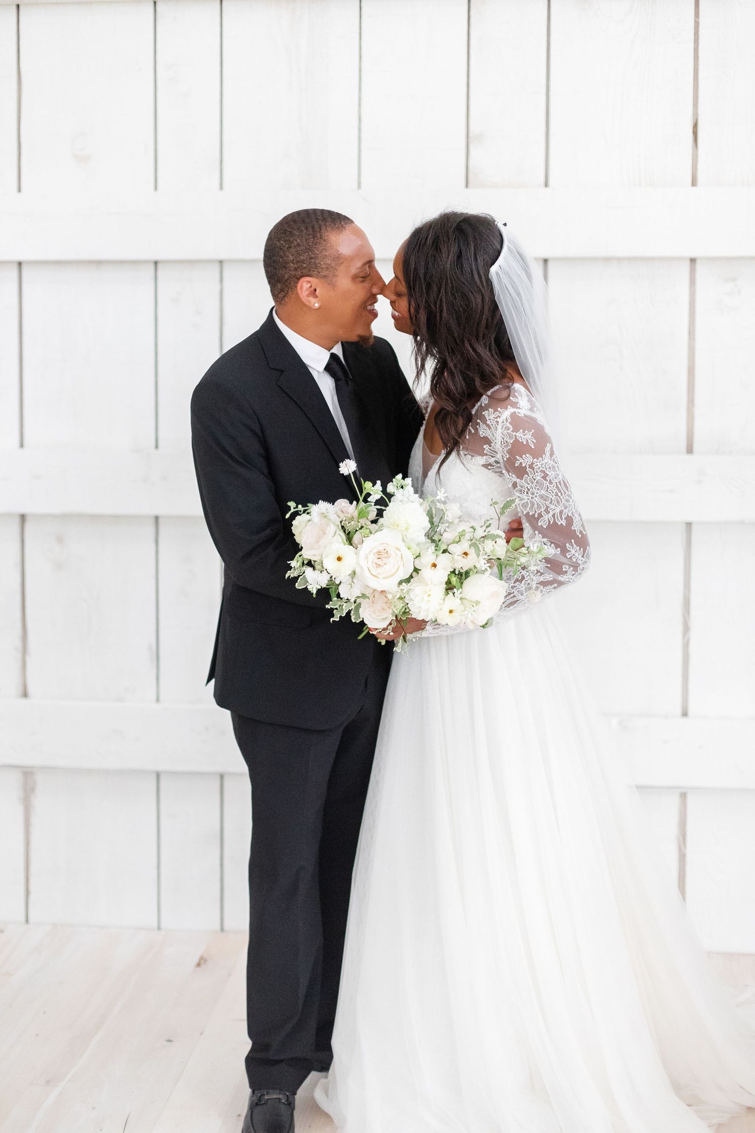Romantic wedding portrait against white wooden wall with elegant white floral bouquet.