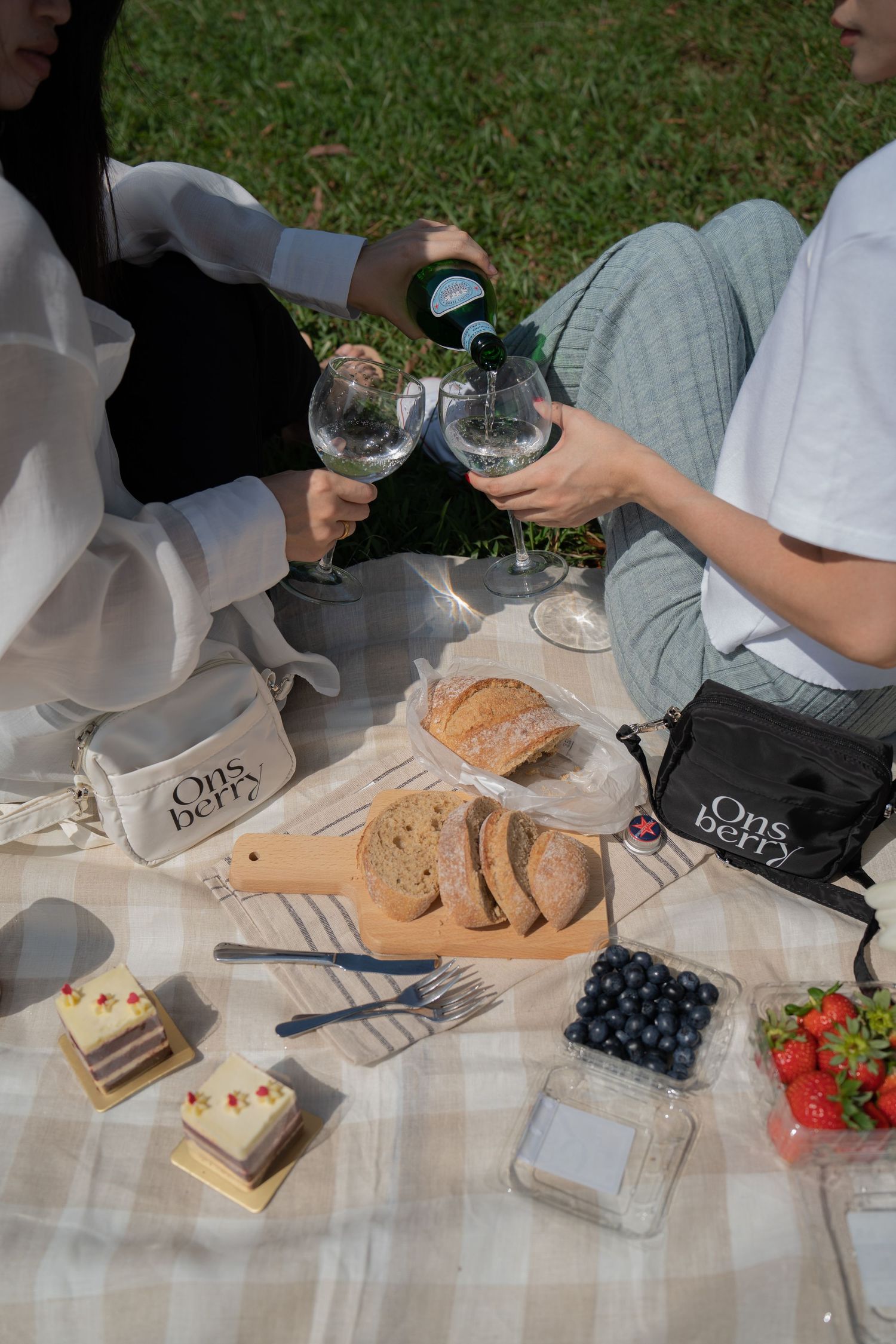 Picnic scene with wine pouring, Ons berry bags, and assorted snacks on white blanket.