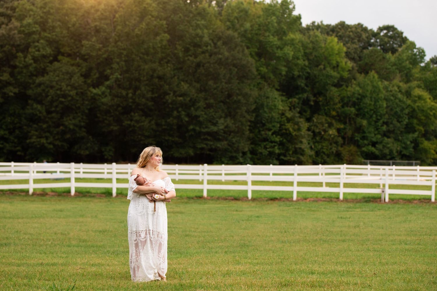 Pastoral maternity scene with white dress against rustic fence and green field.