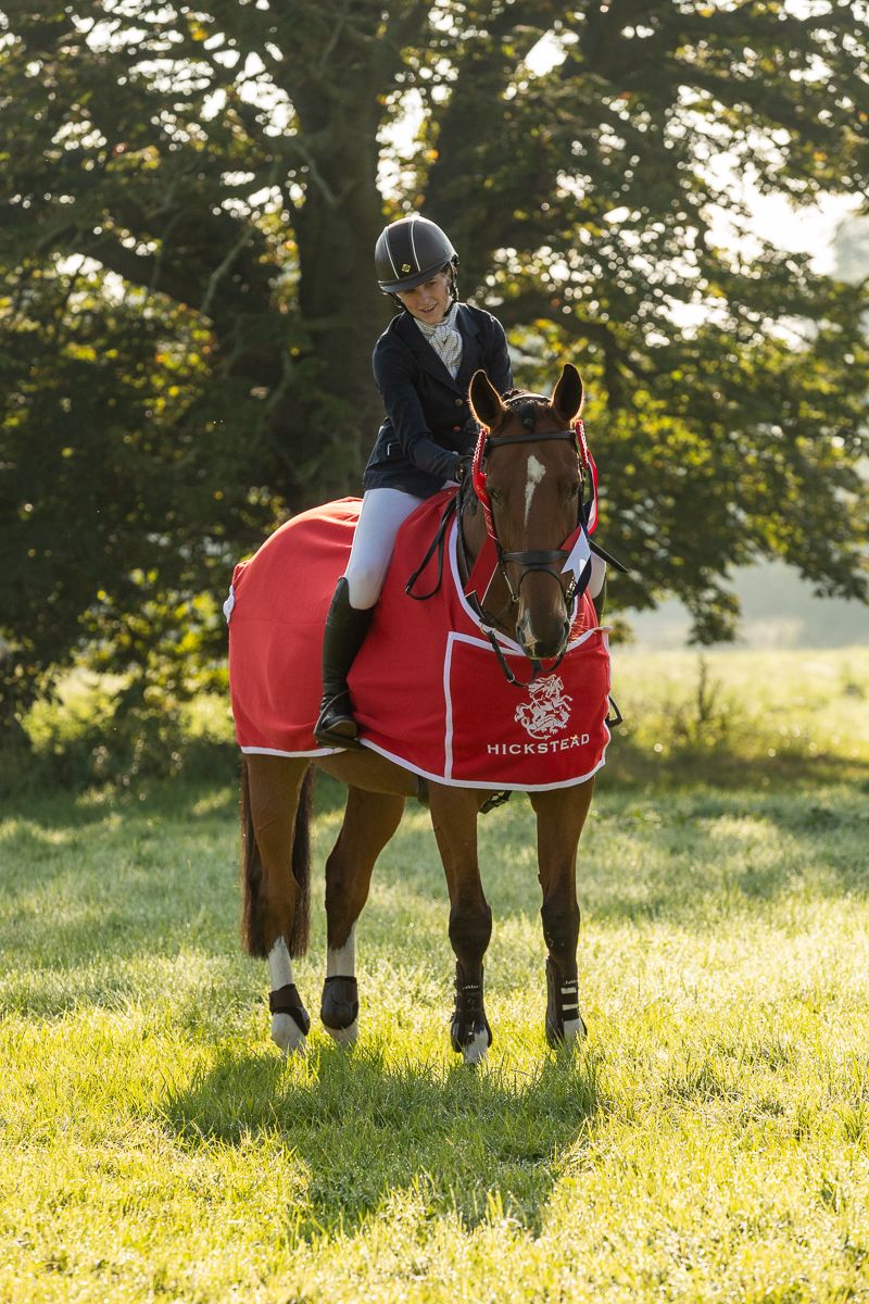 Equestrian rider in competition attire on a bay horse wearing a red show blanket in a sunlit field.