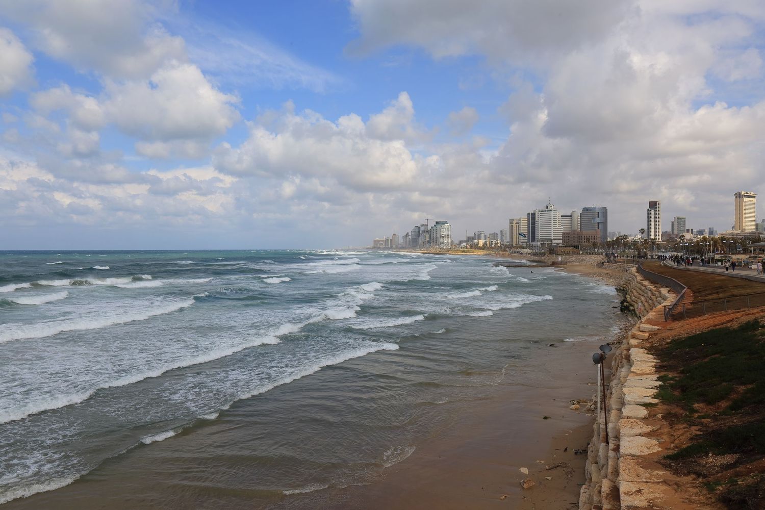 Aerial view of Tel Aviv's coastline with waves crashing on the beach and modern buildings along the Mediterranean shore.