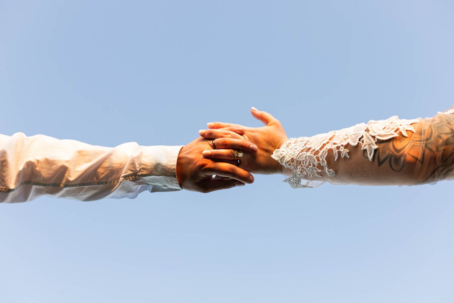 Bride and groom arms holding hands in the center with blue skies above. Detail photograph of rings and dress sleeve