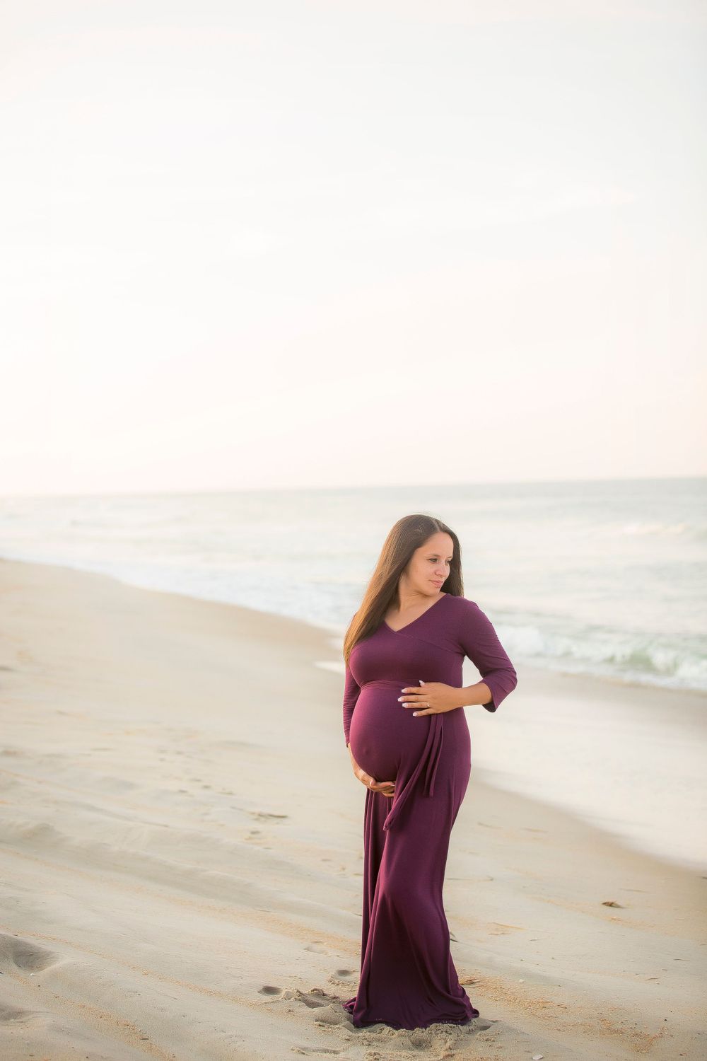 Pregnant mom standing on the beach cradling her belly with a beautiful blue pink sky behind her.