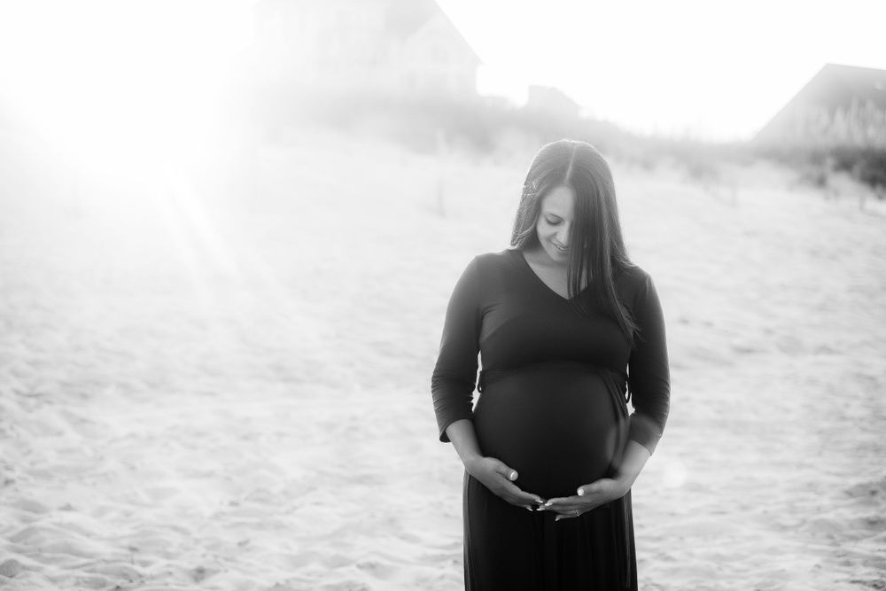 Pregnant mom standing near the sand dunes with sun setting behind her.  She is wearing a burgundy dress.